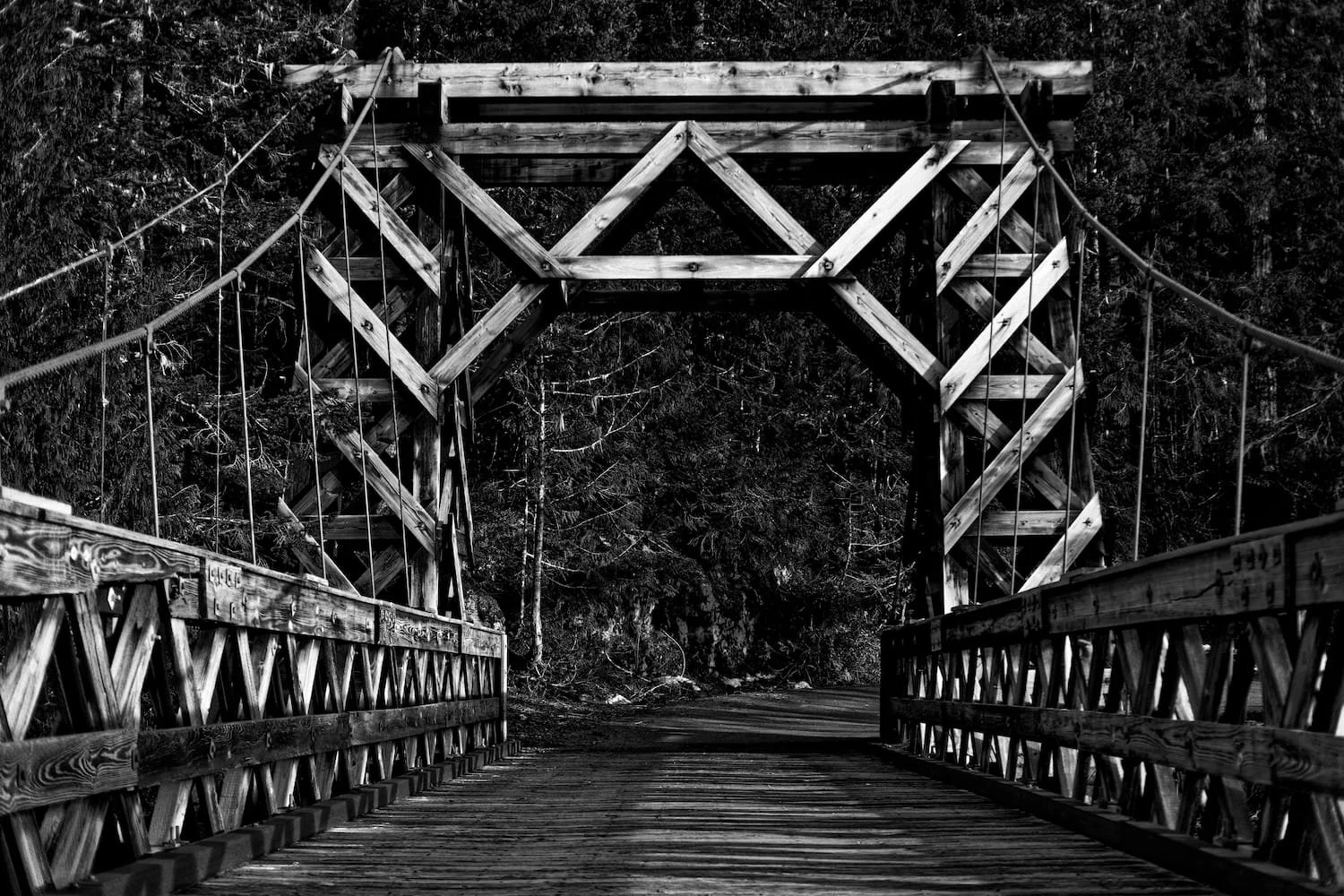 Black and white photo of a wooden suspension bridge with trees in the background.