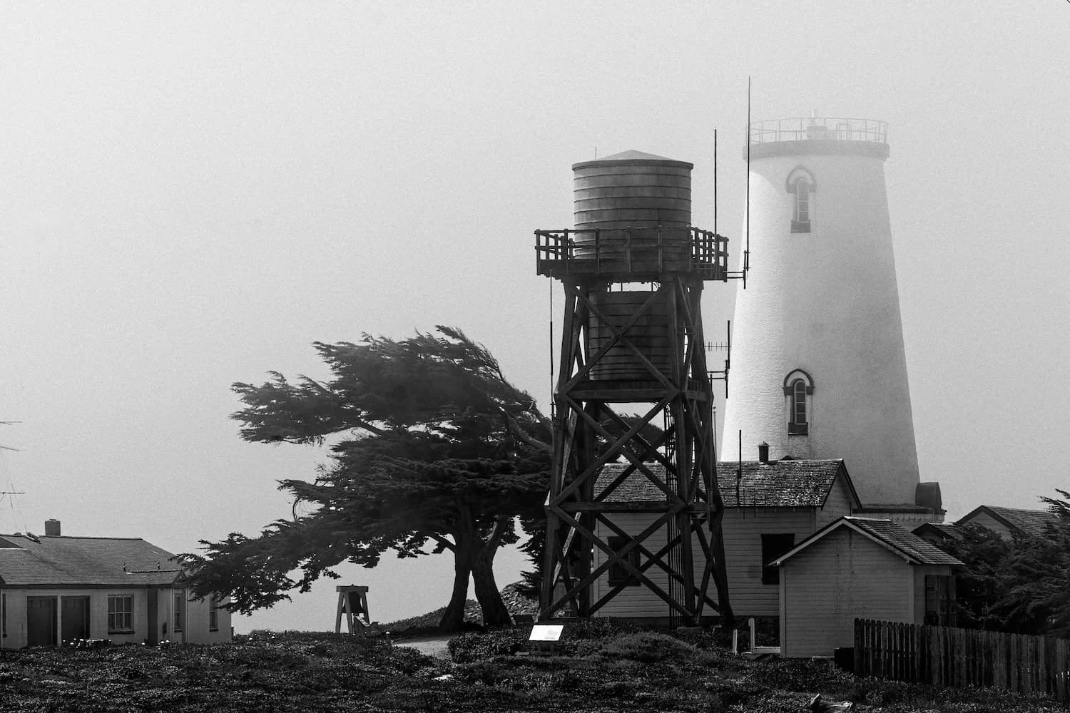 Black and white photo of a lighthouse with a smaller building nearby, a water tower on a wooden structure, and trees in the background.