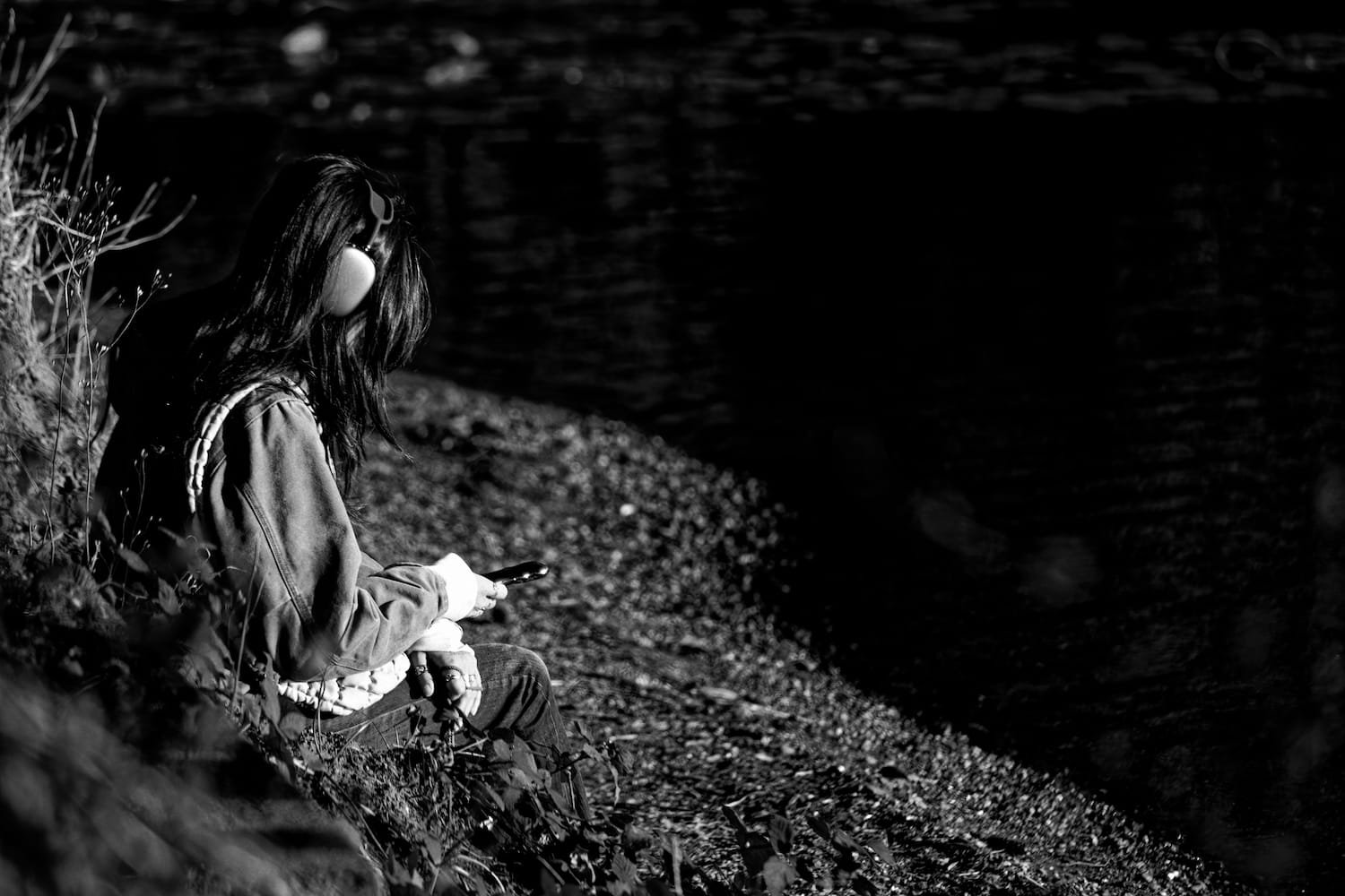 A person with long hair wearing headphones, a hoodie, and jeans sitting on a rocky shoreline, looking at a phone, with a body of water in the background, in black and white.