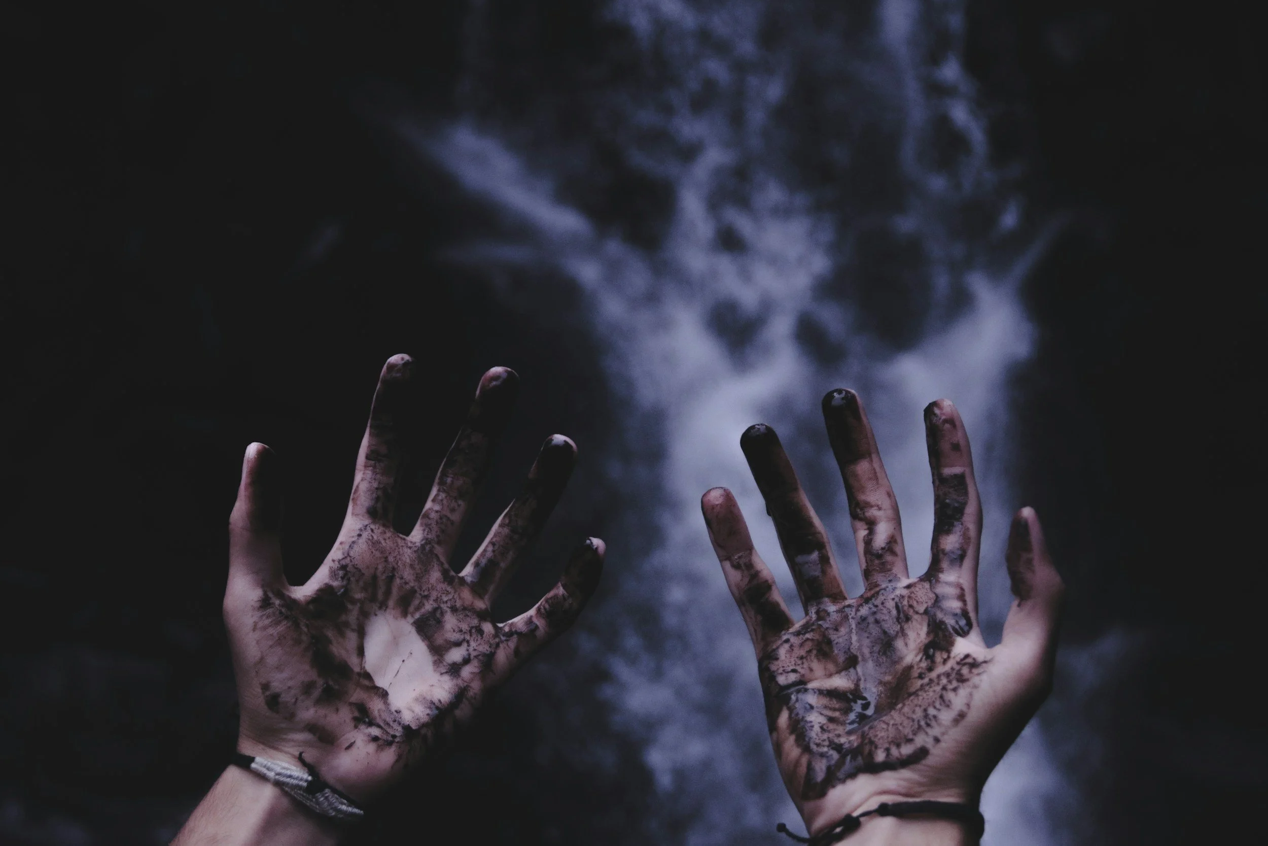 Two hands with dirty, muddy fingers reaching toward a dark, misty background.