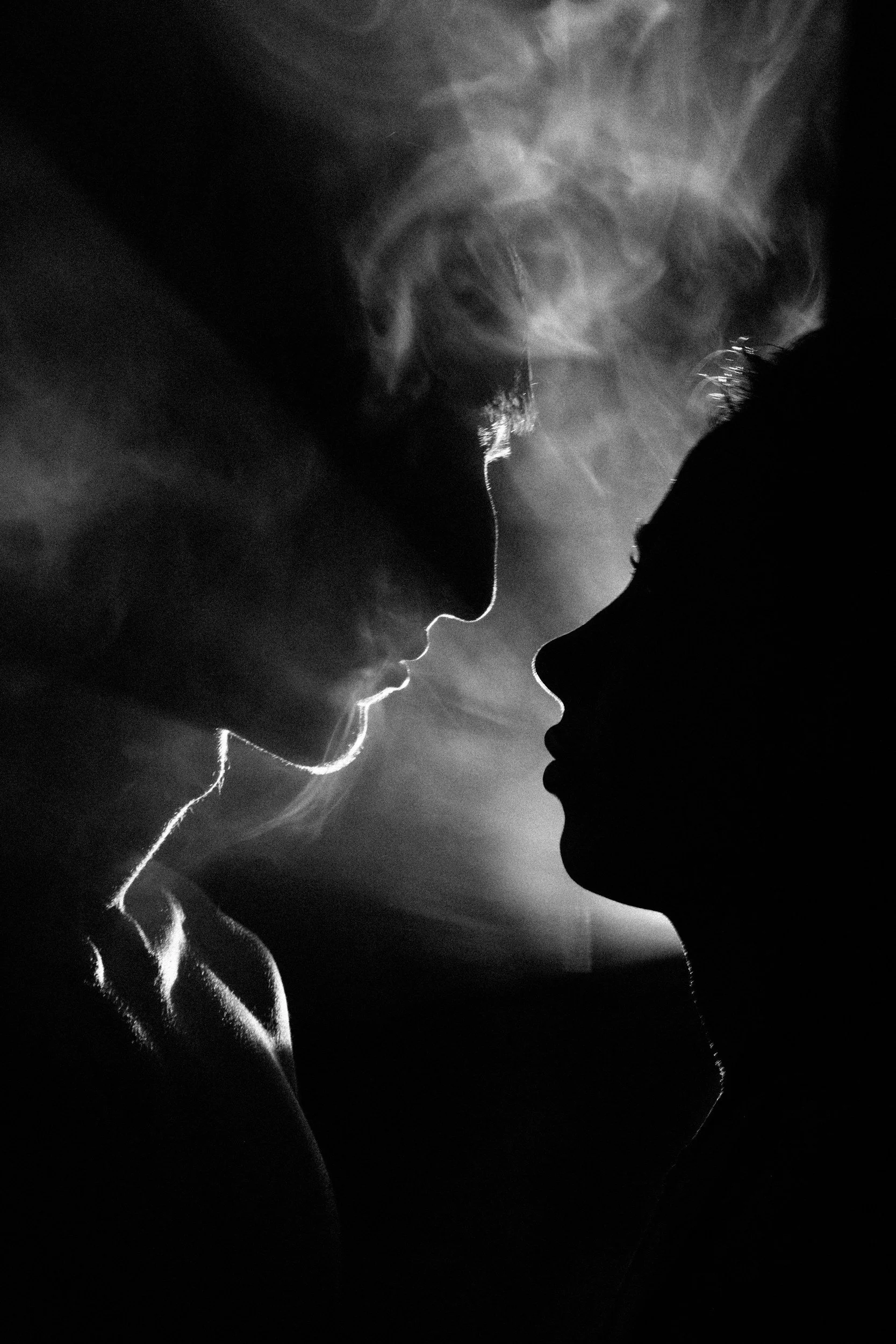 Silhouettes of two people close to each other, with light highlighting their profiles and smoke around their faces.