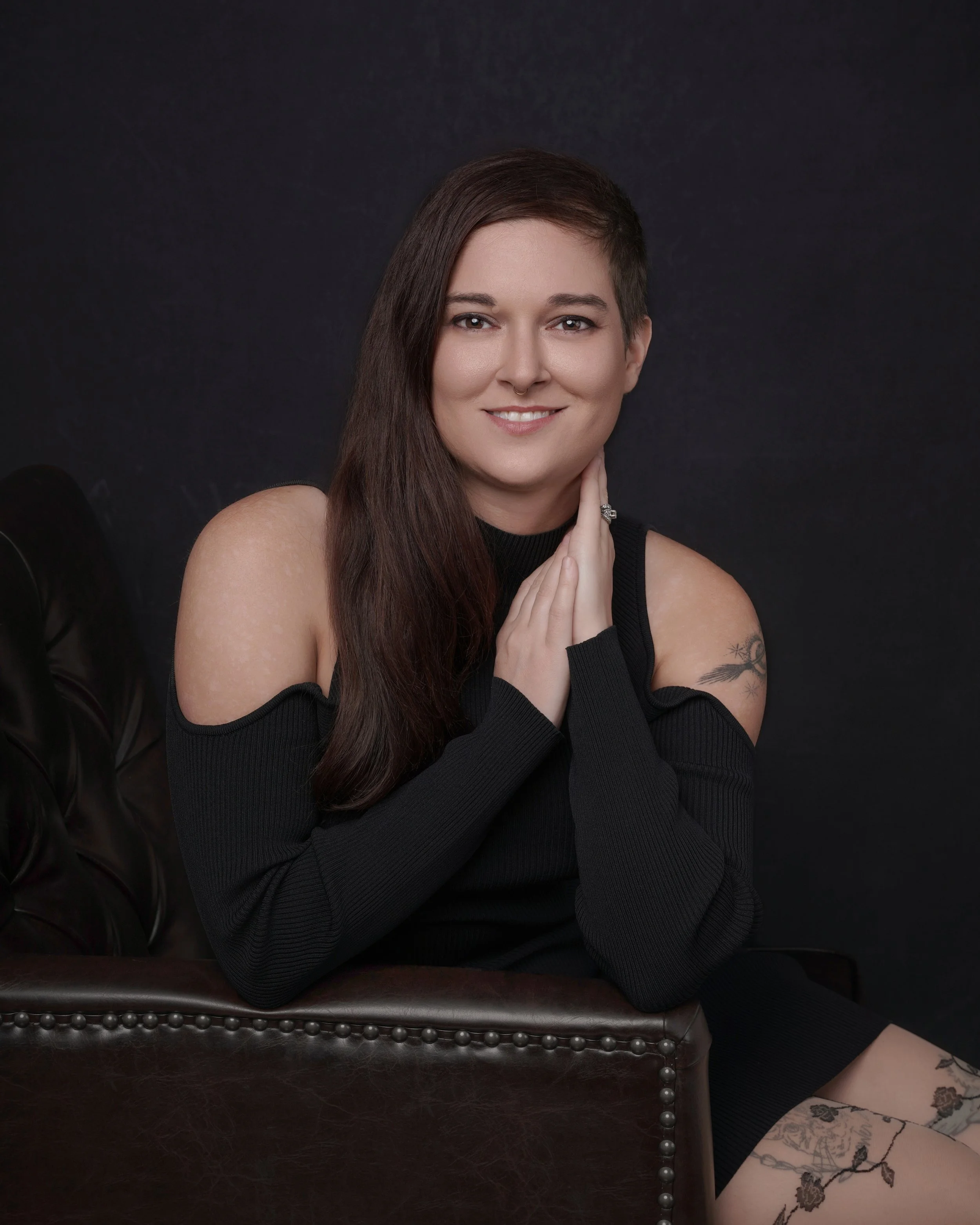 A young woman with long dark hair smiling and sitting on a dark leather chair against a black background. She is wearing a black cold-shoulder top and patterned tights. author j.f. lane