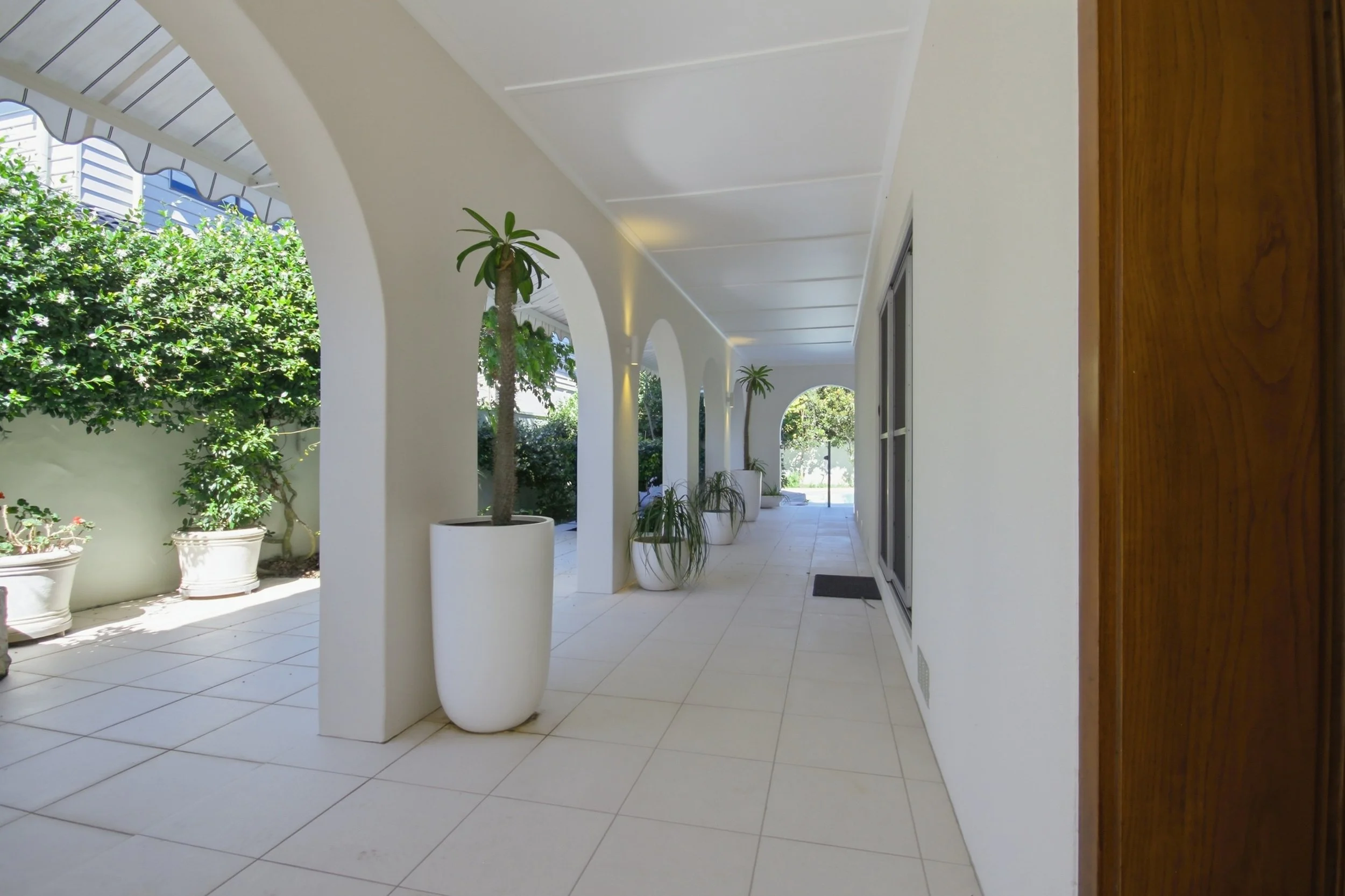Covered outdoor walkway with white tiled floor, white walls, arched openings, potted plants, and greenery outside.