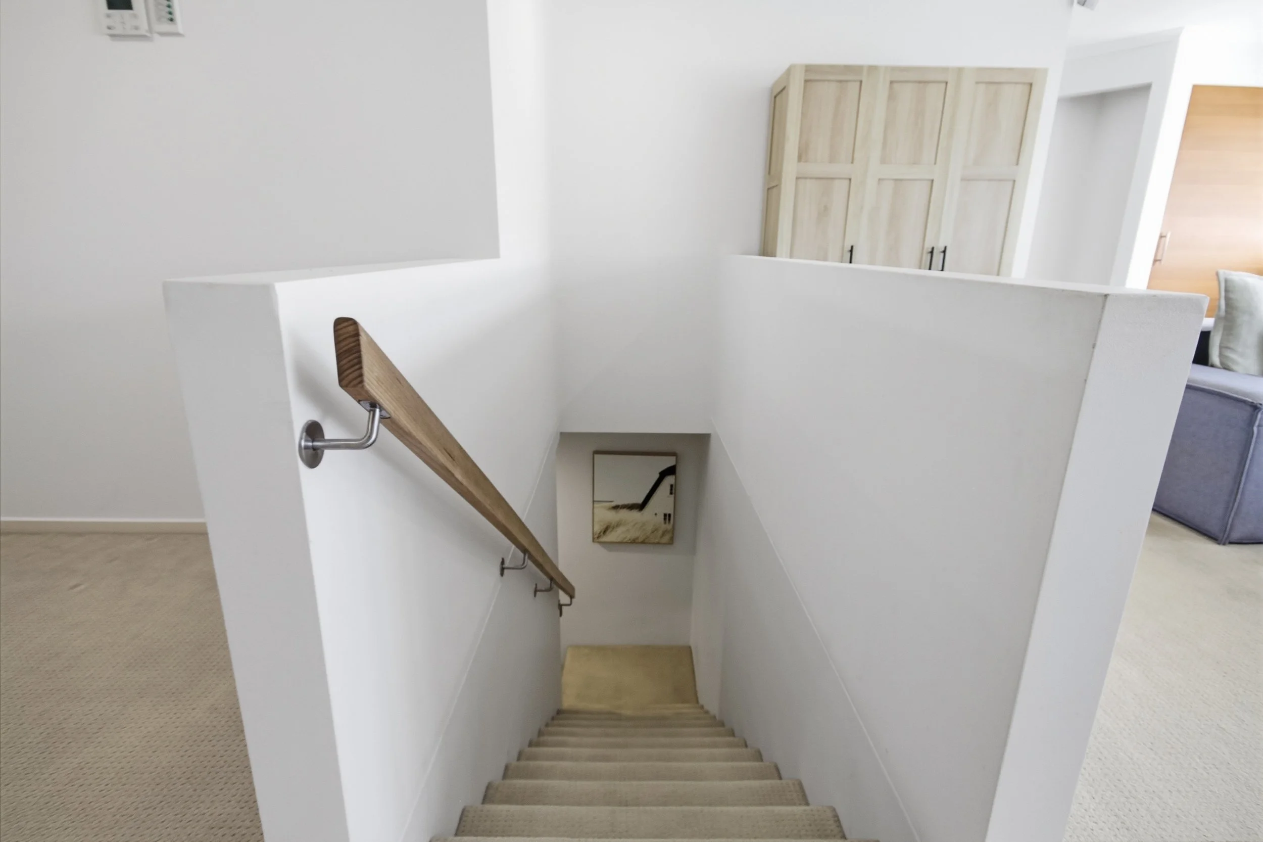 Top view of a staircase with beige carpeted steps, white walls, a wooden handrail on the left, leading down to a landing with a framed picture, and a storage cabinet in the background.