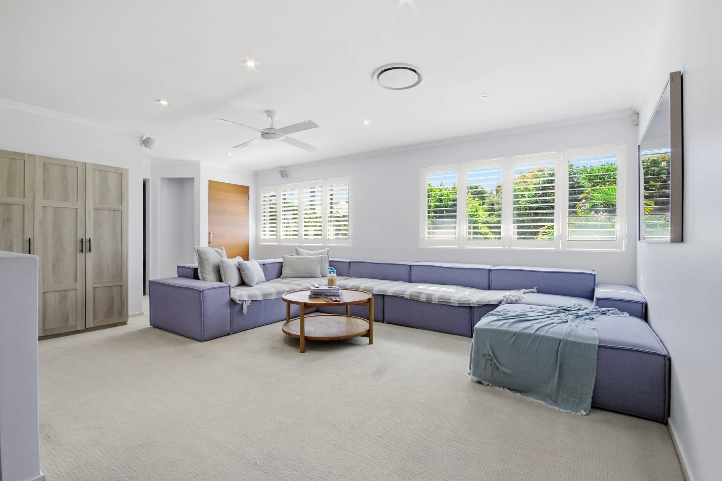 Bright living room with large L-shaped purple sofa, beige carpet, white walls, ceiling fan, multiple windows with white plantation shutters, wall-mounted TV, and minimal decor.