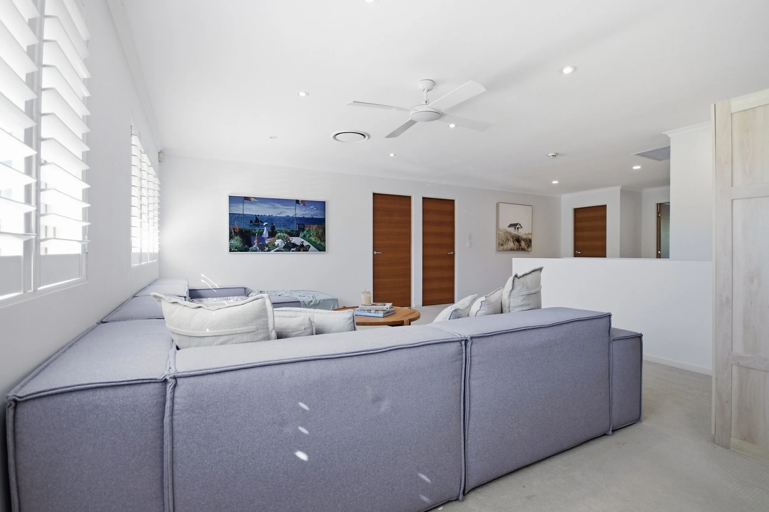 Modern living room with gray sectional sofa, white pillows, wooden coffee table with books and candles, wall art, white walls, ceiling fan, and wooden doors.