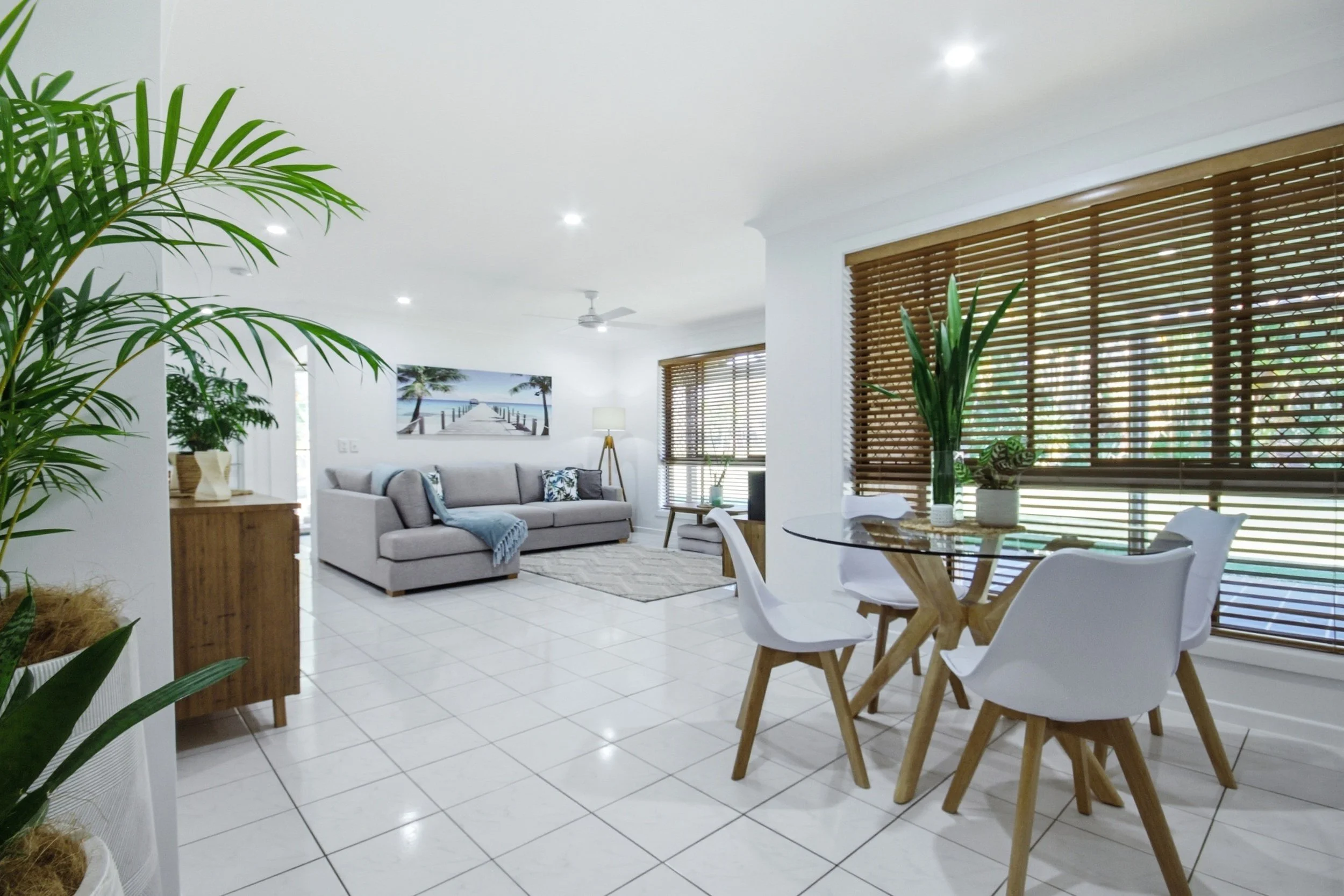 Bright living room and dining area with white tiled floors, wooden blinds, a gray sofa, round glass-top dining table with white chairs, and many green plants.
