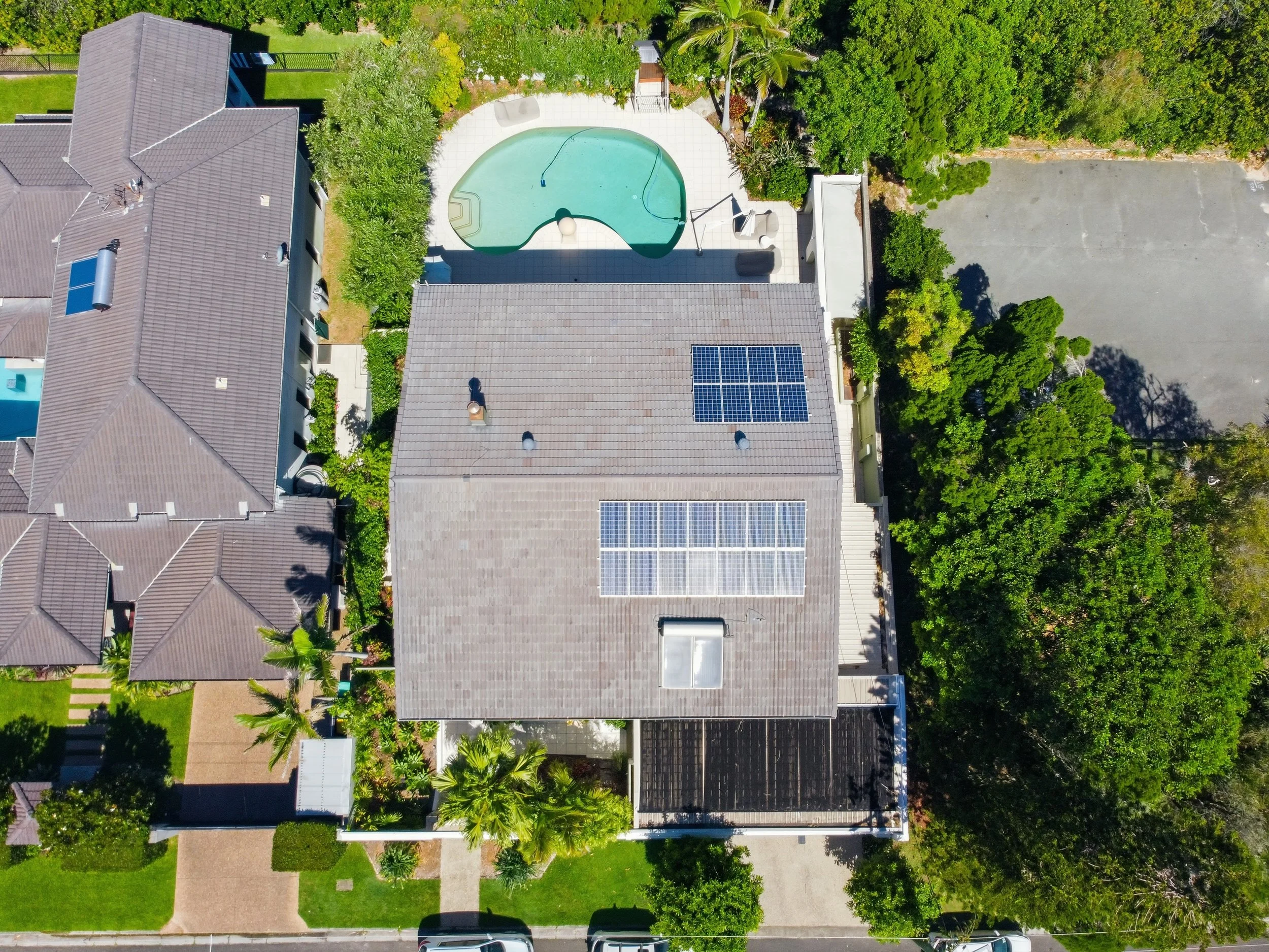 Aerial view of a backyard with a kidney-shaped swimming pool, surrounded by a patio and greenery. There are solar panels on the roof of the house, and neighboring houses are visible. There's a gravel parking lot, trees, and a sidewalk along the front