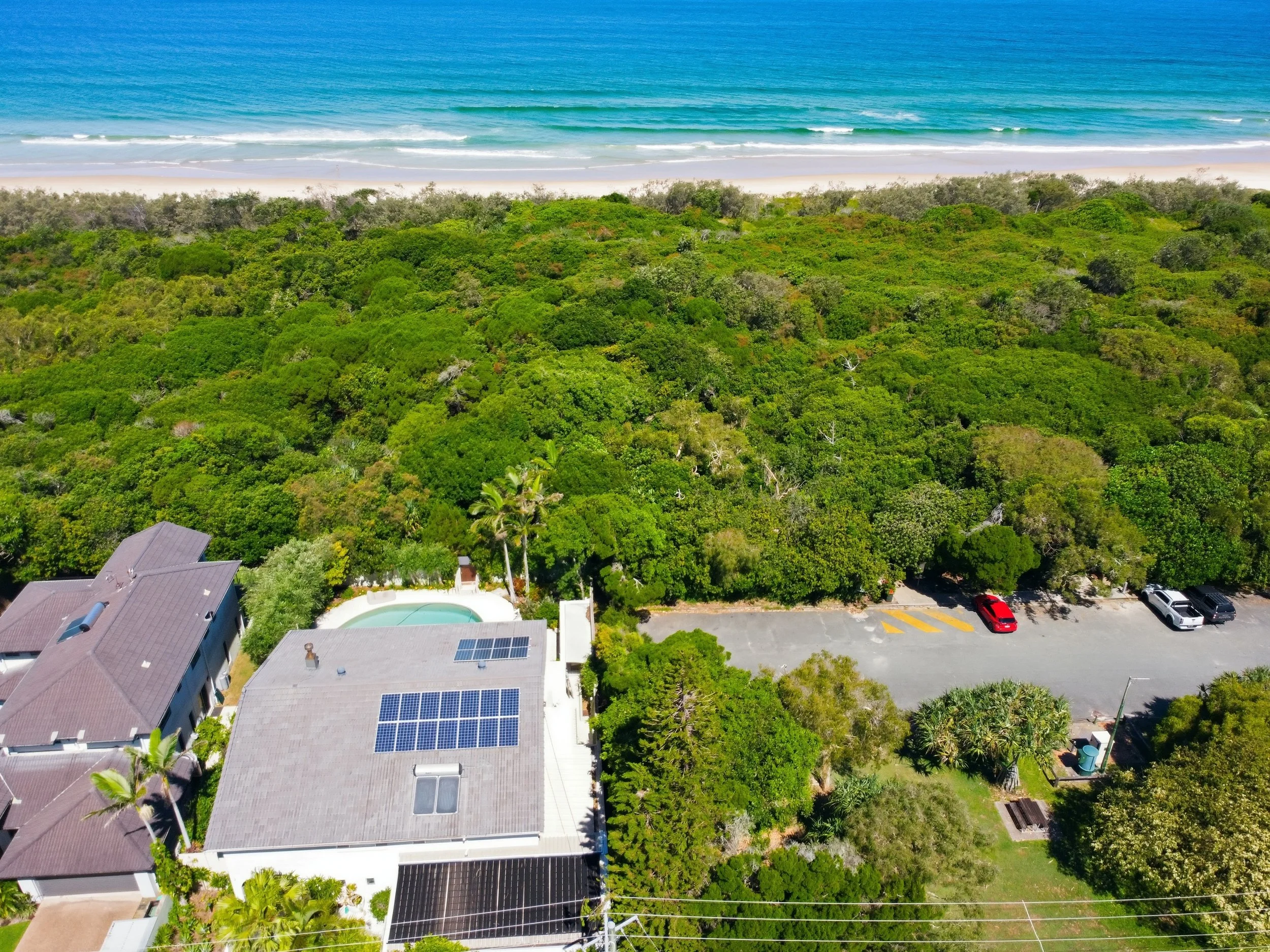 Aerial view of a beachfront area with green trees, residential buildings with solar panels, a swimming pool, and a parking lot with a few cars, with the ocean in the background.