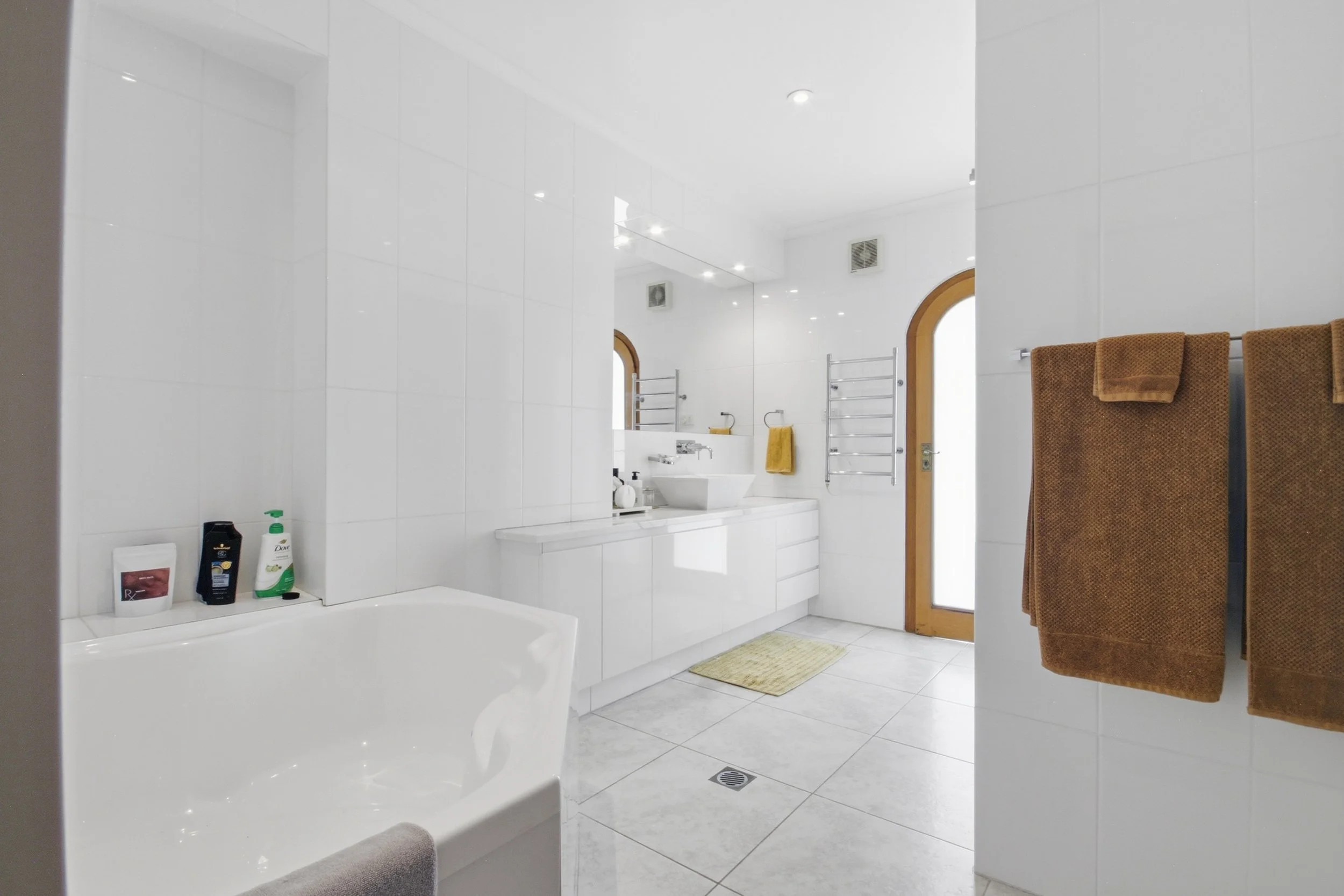Bright bathroom with white tiled walls and floor, a bathtub, a white vanity with a sink, large mirror, and brown towels hanging on a wall rack.