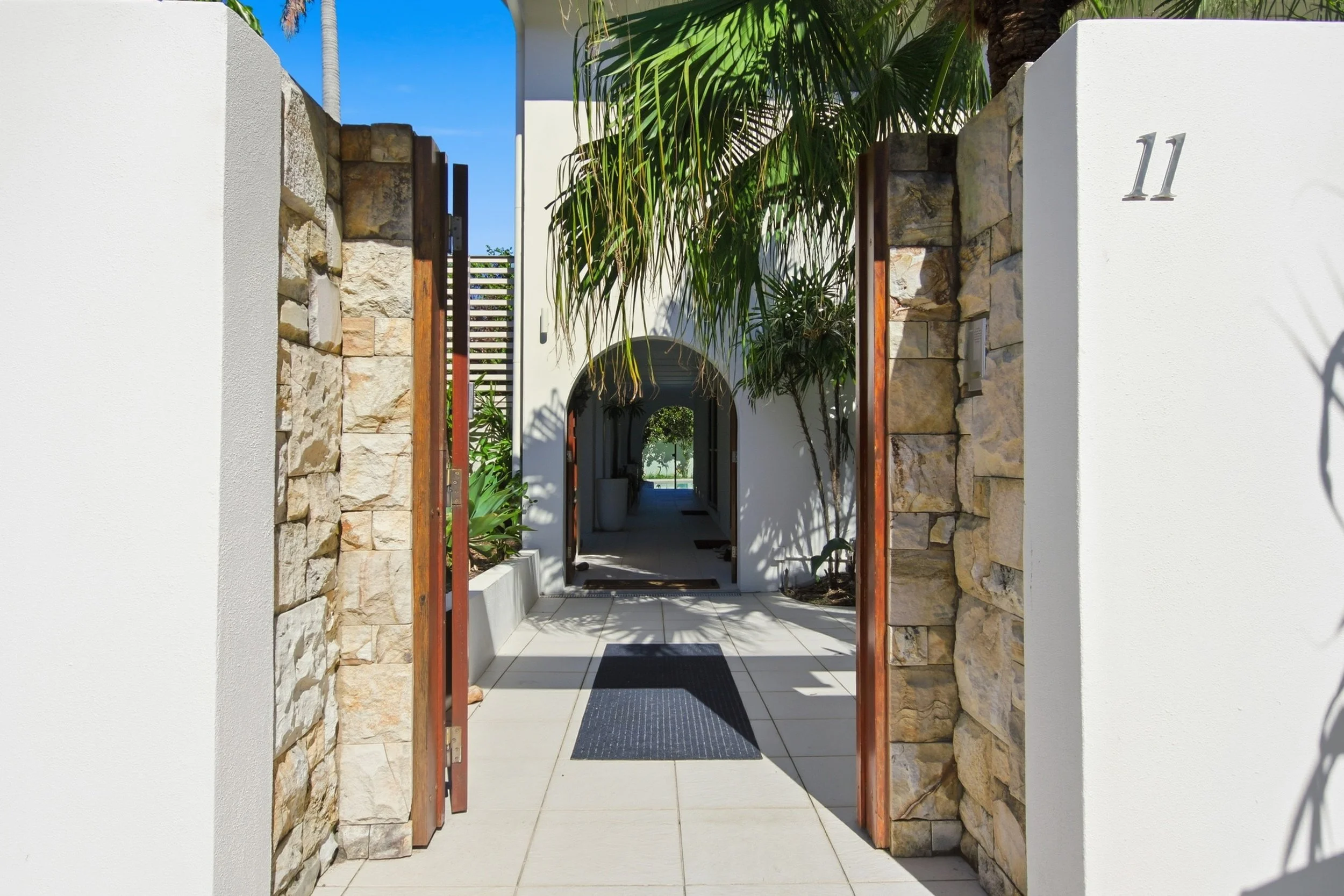 Modern entrance with stone and white walls, open gate, tropical plants, arches, and pathway leading to a backyard.