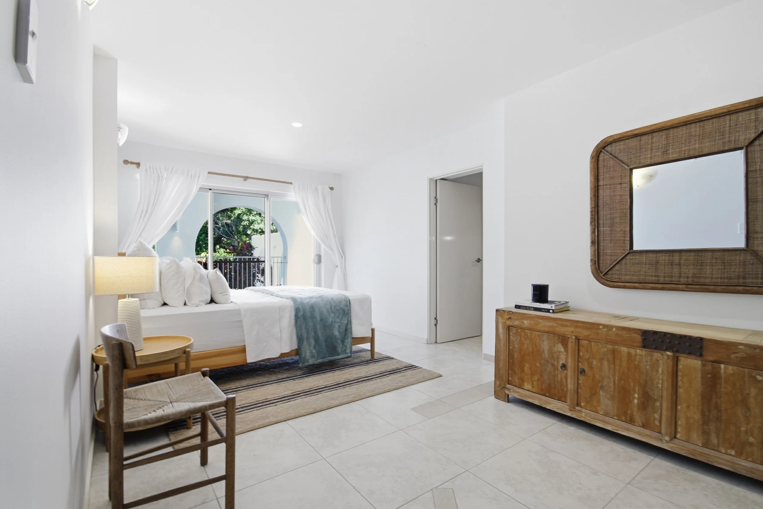 Bright bedroom with a large bed, white bedding, and four pillows, next to a sliding door opening to a balcony with greenery outside. There is a wooden chair with a woven seat and a side table with a lamp next to the bed. On the right, a wooden sideboard and a mirror with a wooden frame are visible.