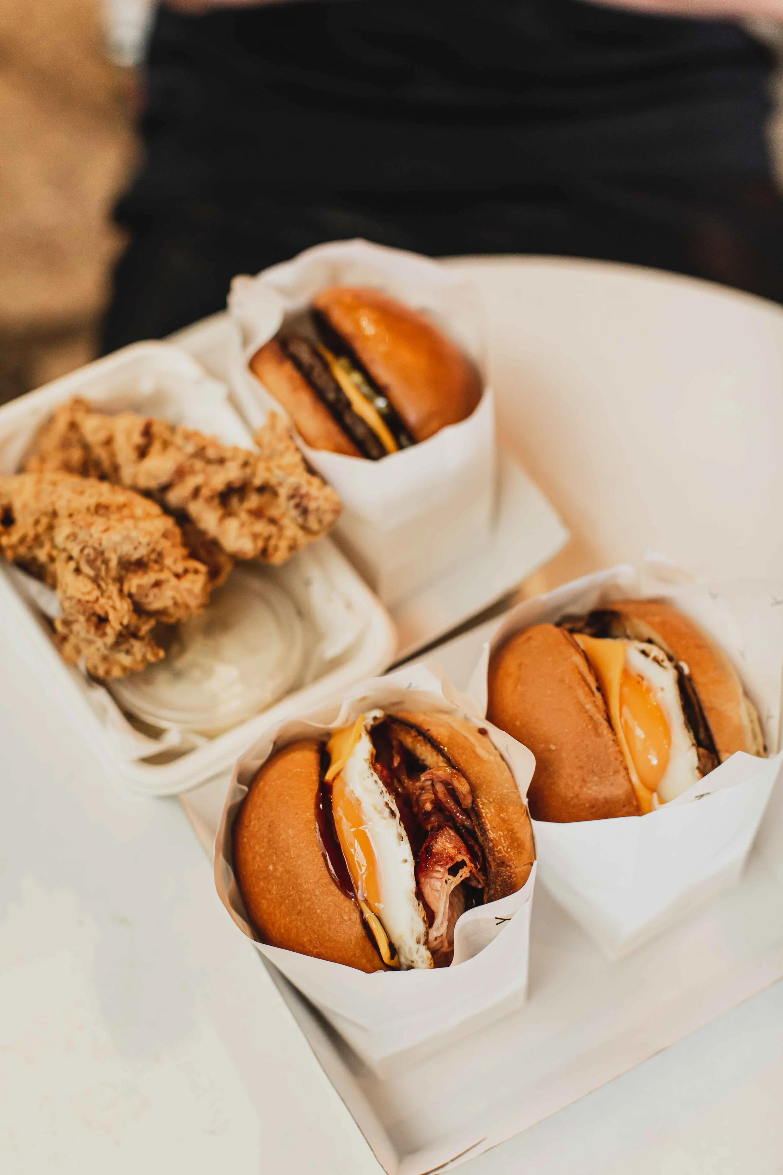 Three breakfast rolls with bread, cheese, and meat, wrapped in white paper, placed in a white paper tray with a serve of fried chicken.
