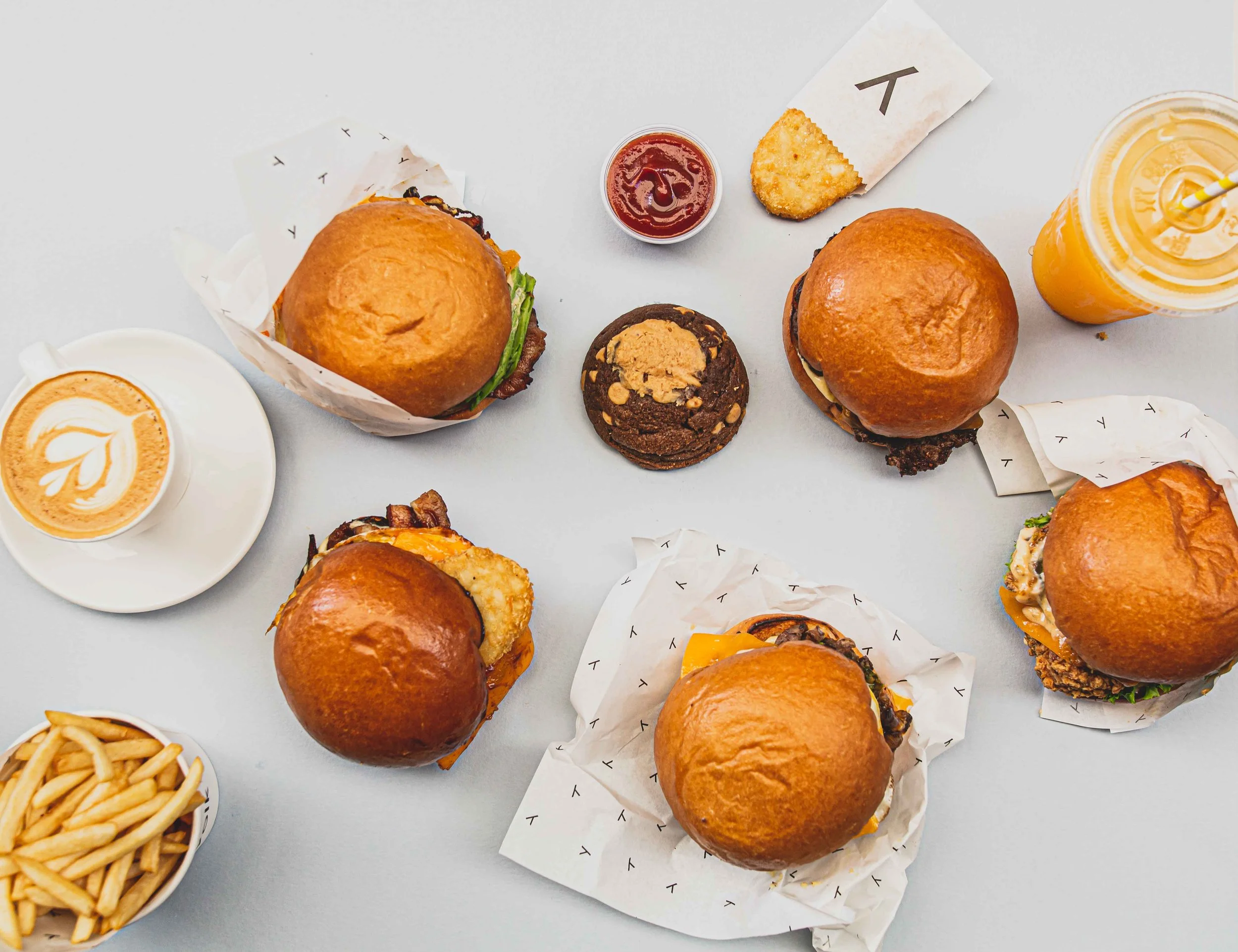 Overhead view of three hamburgers, a cup of coffee with latte art, a cookie, a hash brown, a sandwich with a straw in a drink, and a bowl of French fries on a white surface.