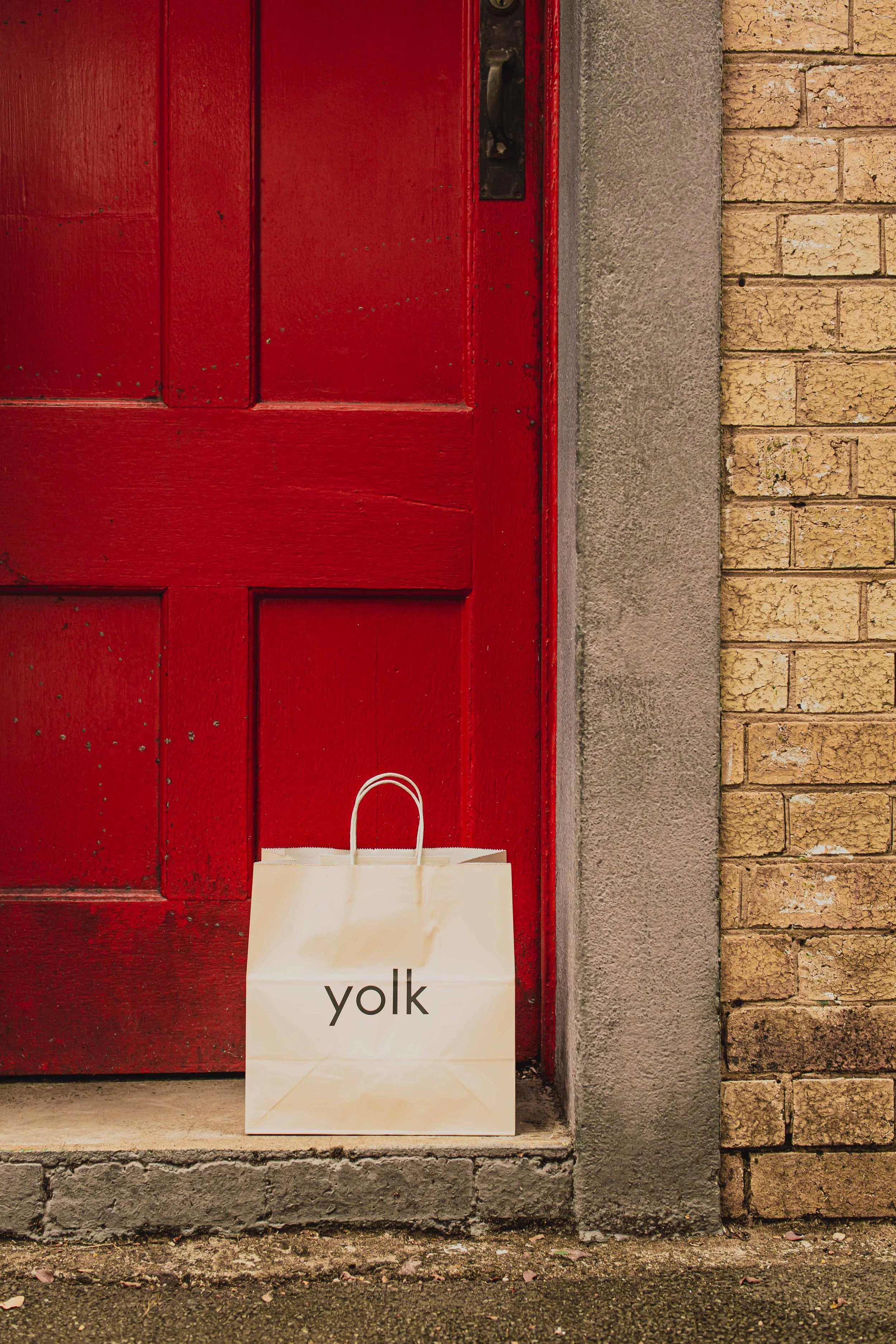 Cream-colored paper takeaway bag with the word 'yolk' printed on it, placed in front of a red door with a black handle, adjacent to a brick wall.