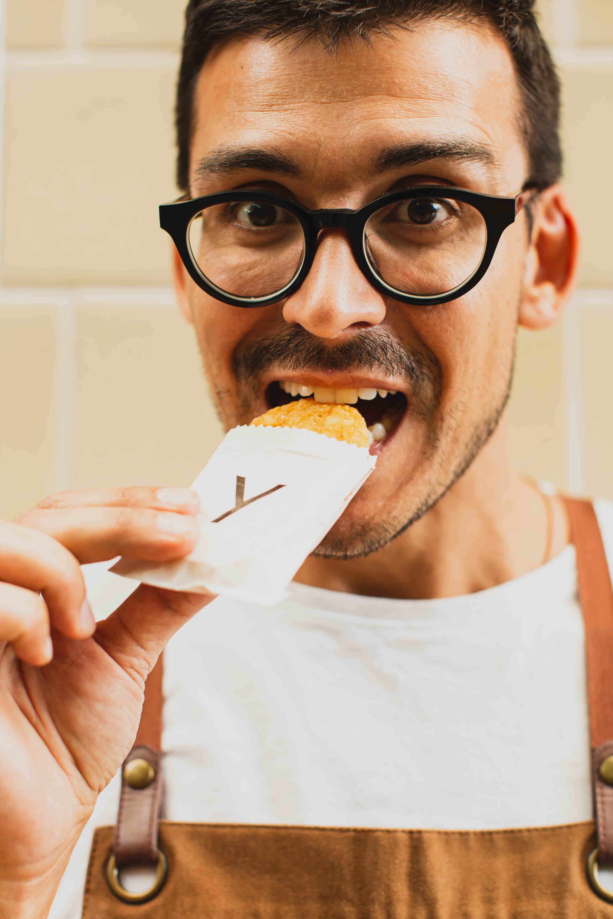 A man with black glasses and a mustache takes a bite of a fried hash brown wrapped in white paper. He is wearing a white shirt and brown overalls.