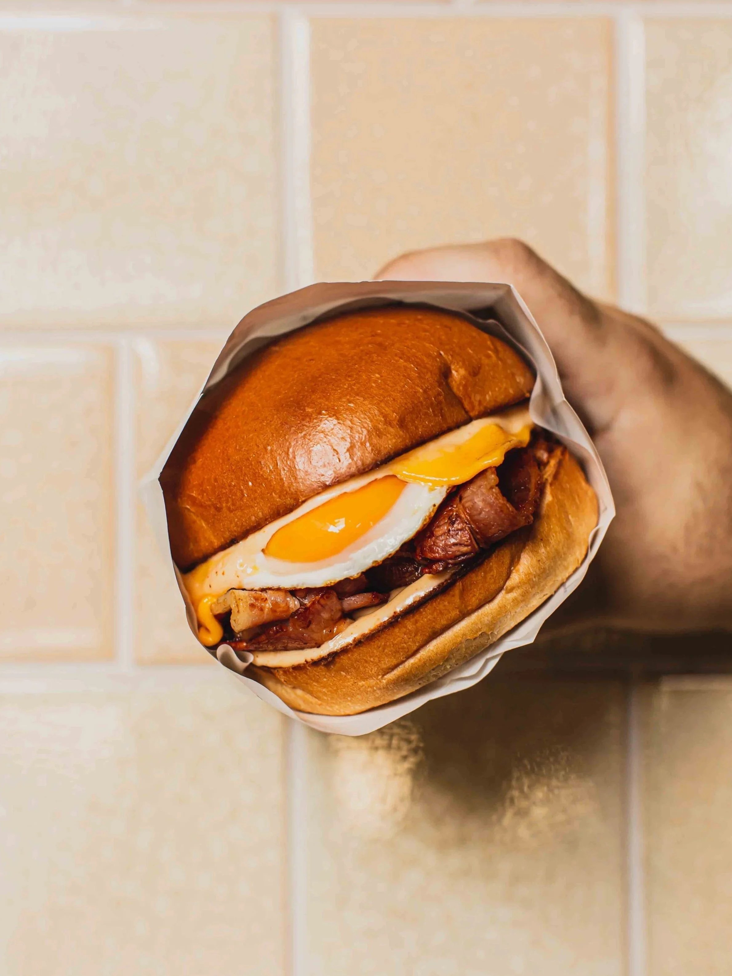Close-up of a bacon and egg roll with a fried egg, melted cheese, bacon, and a toasted bun, being held above a tiled kitchen or restaurant wall.