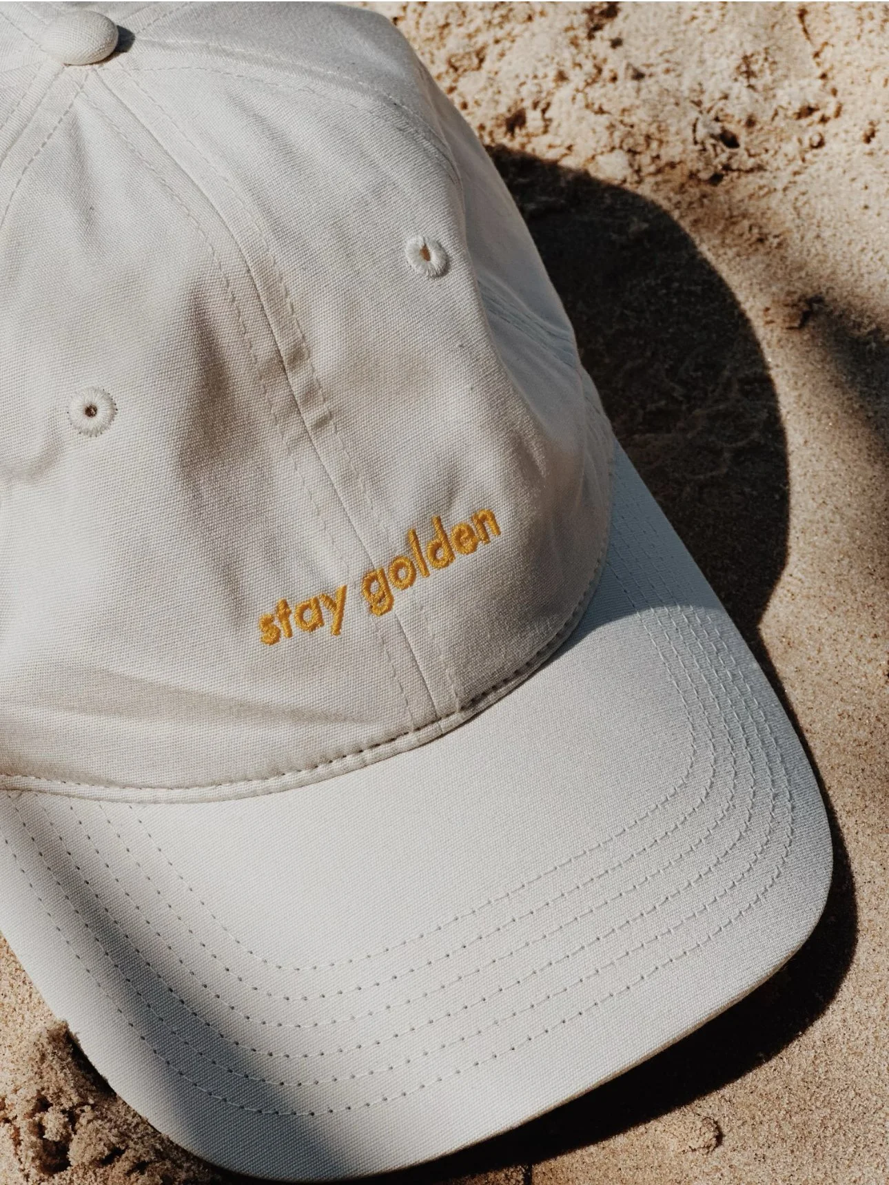 Close-up of a white baseball cap with orange embroidered text that reads 'stay golden,' resting on sand.