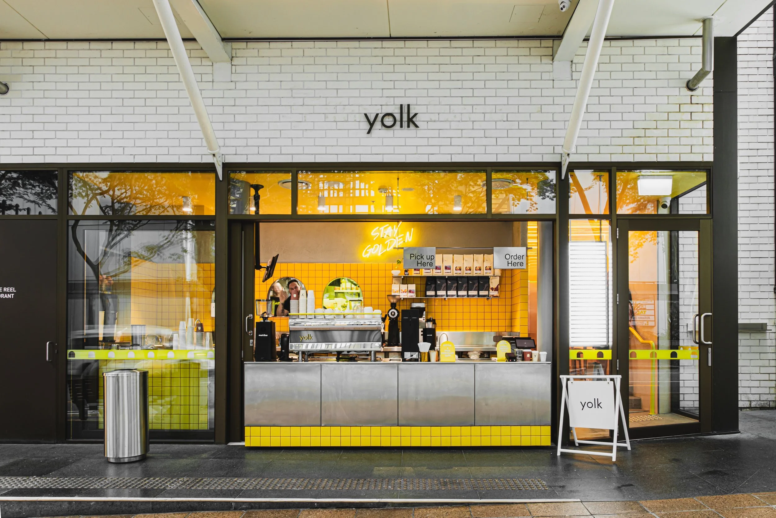 Front view of a bright yellow and black coffee shop named 'yolk' with large glass windows and a door. Inside, a barista is preparing drinks behind the counter, with a neon sign that reads 'Stay Golden.' The shop has a modern design with yellow tiles and warm lighting.