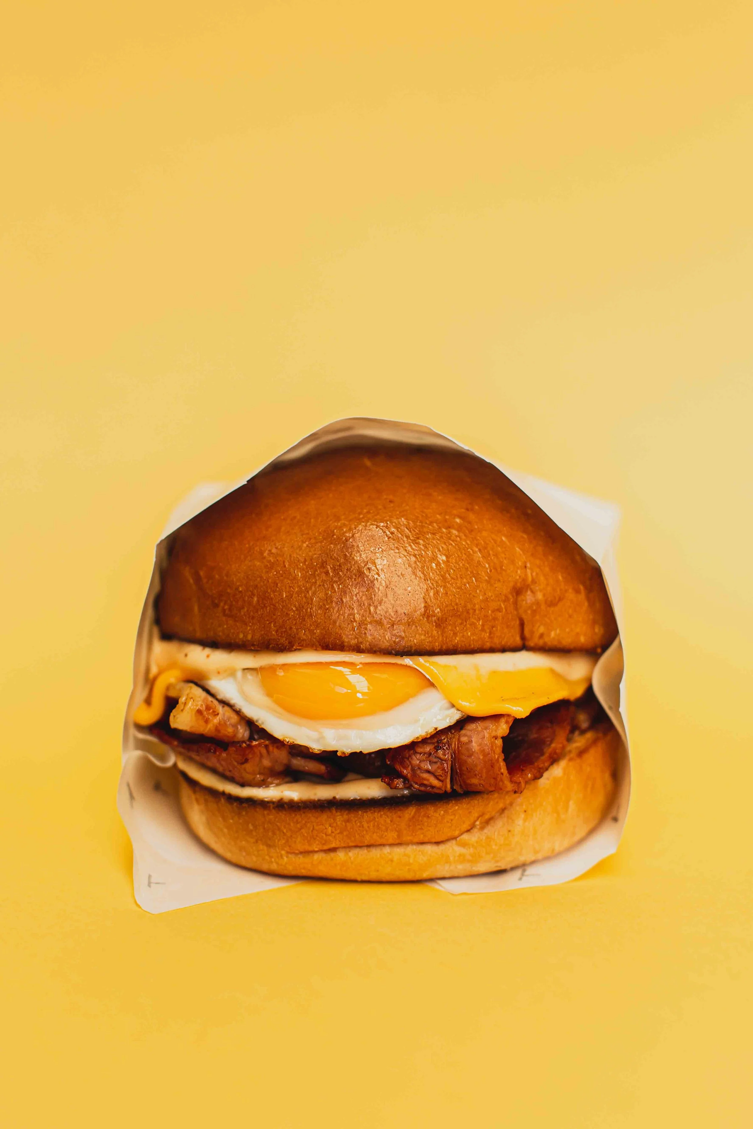 Close-up of a bacon and egg roll with a toasted bun, fried chicken, a sunny side up egg, melted cheese, and bacon, against a yellow background.