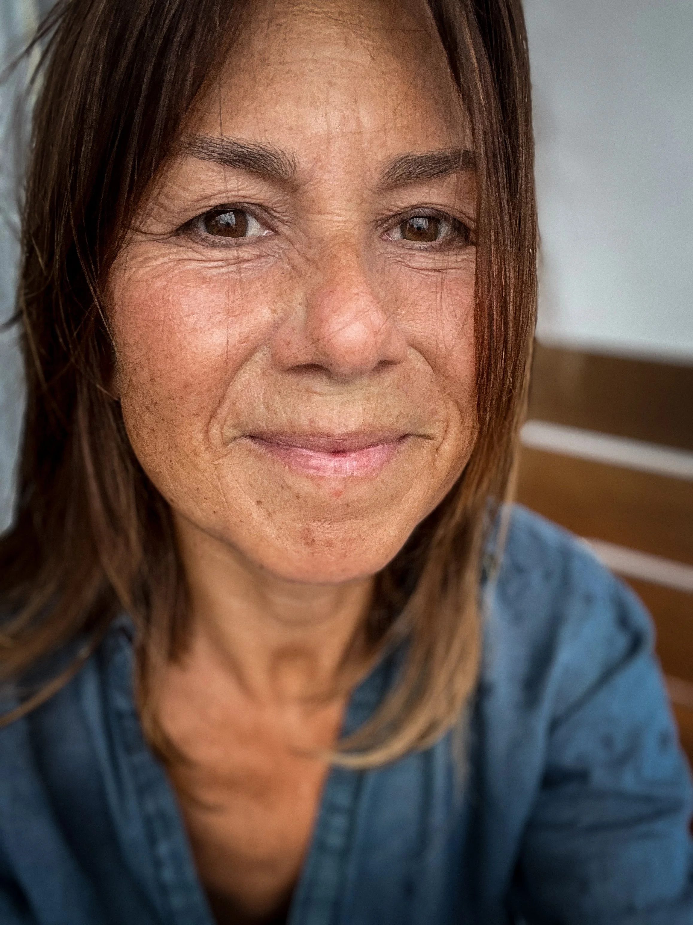 A woman with medium-length brown hair, brown eyes, and light skin, smiling slightly, wearing a denim jacket, sitting on a wooden bench.