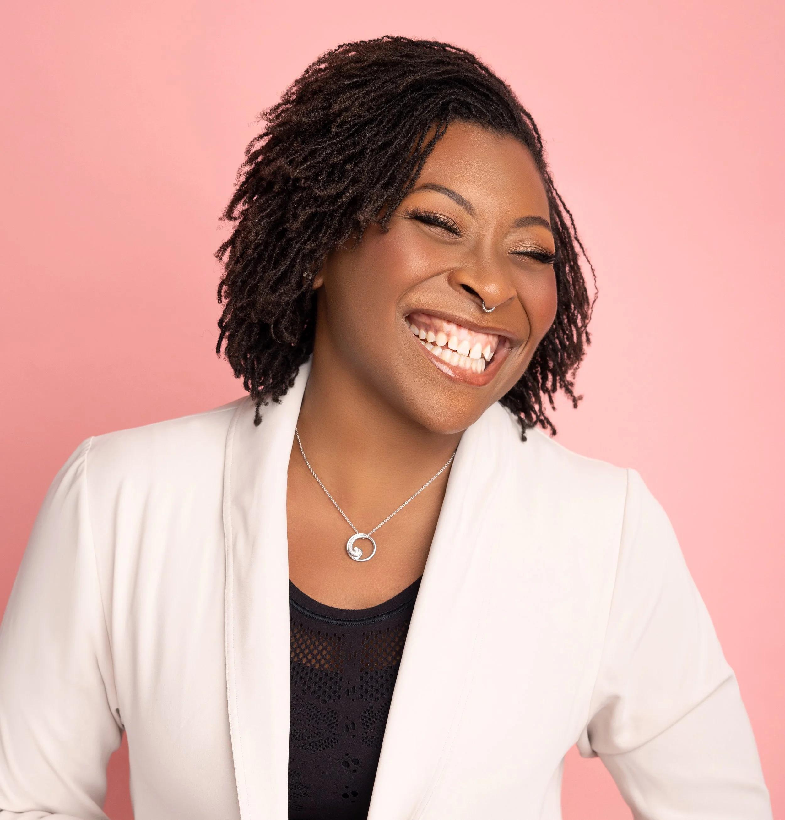 Christina poses and smiles for a professional photo with a pink backdrop. She has styled her dreadlocks and is wearing an off-white blazer and black blouse underneath.