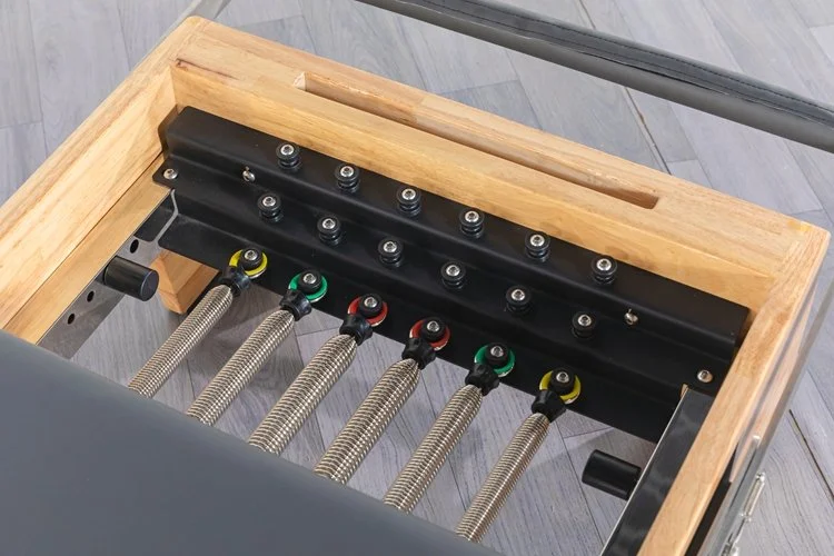 Close-up of a Pilates reformer with metal springs, color-coded for resistance, attached to a wooden frame.