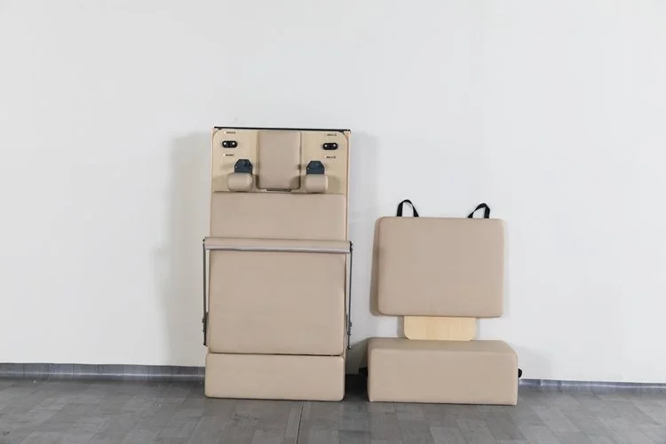Hospital baby changing station with padded seat and two changing surfaces, adjacent to a padded backrest on a plain white wall.