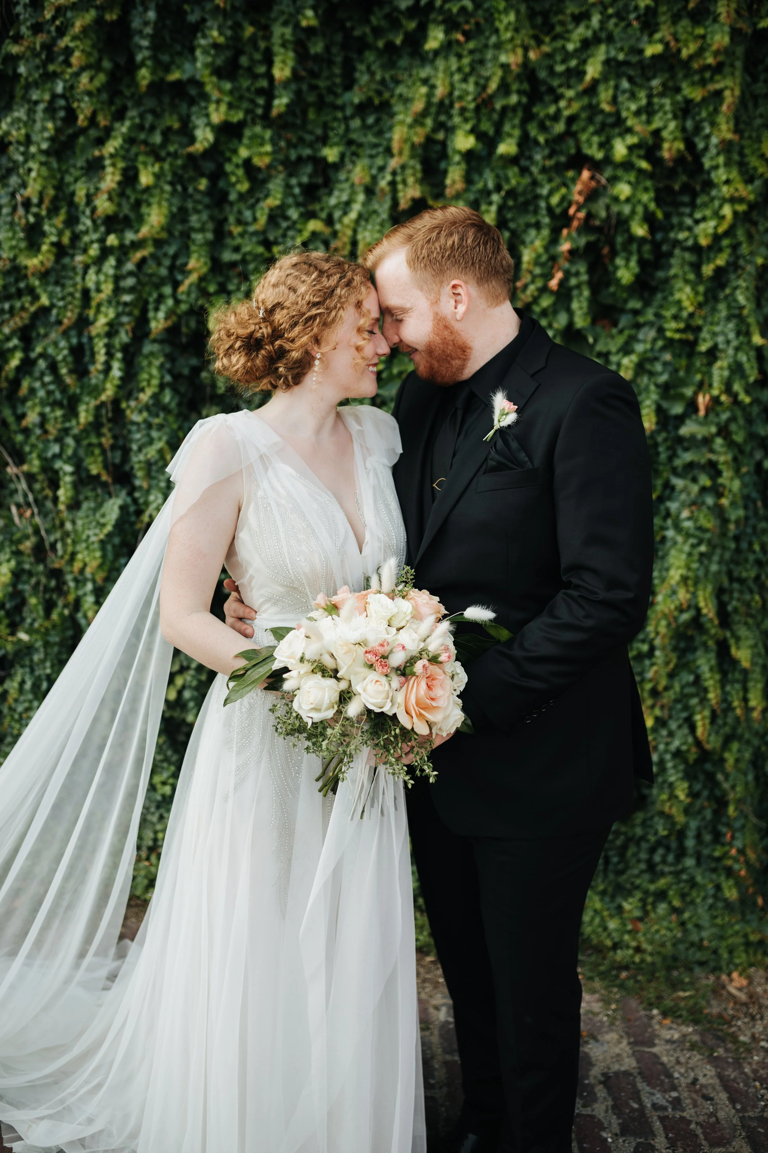 A smiling bride and groom in wedding attire posing outdoors with a green leafy background, holding a bouquet of flowers.