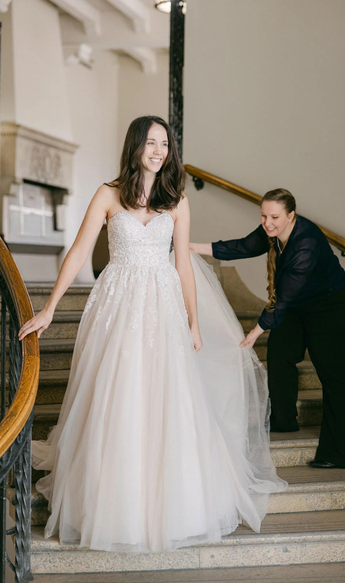 A woman in a white wedding dress standing on a staircase, smiling, as another woman adjusts the dress.