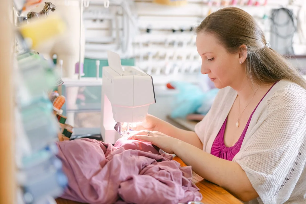 A woman sewing pink fabric on a sewing machine in a craft room or workshop, with shelves and various supplies in the background.
