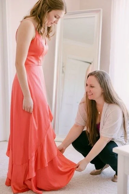 A woman in a coral dress helps another woman try on the dress, both smiling, in a brightly lit room.