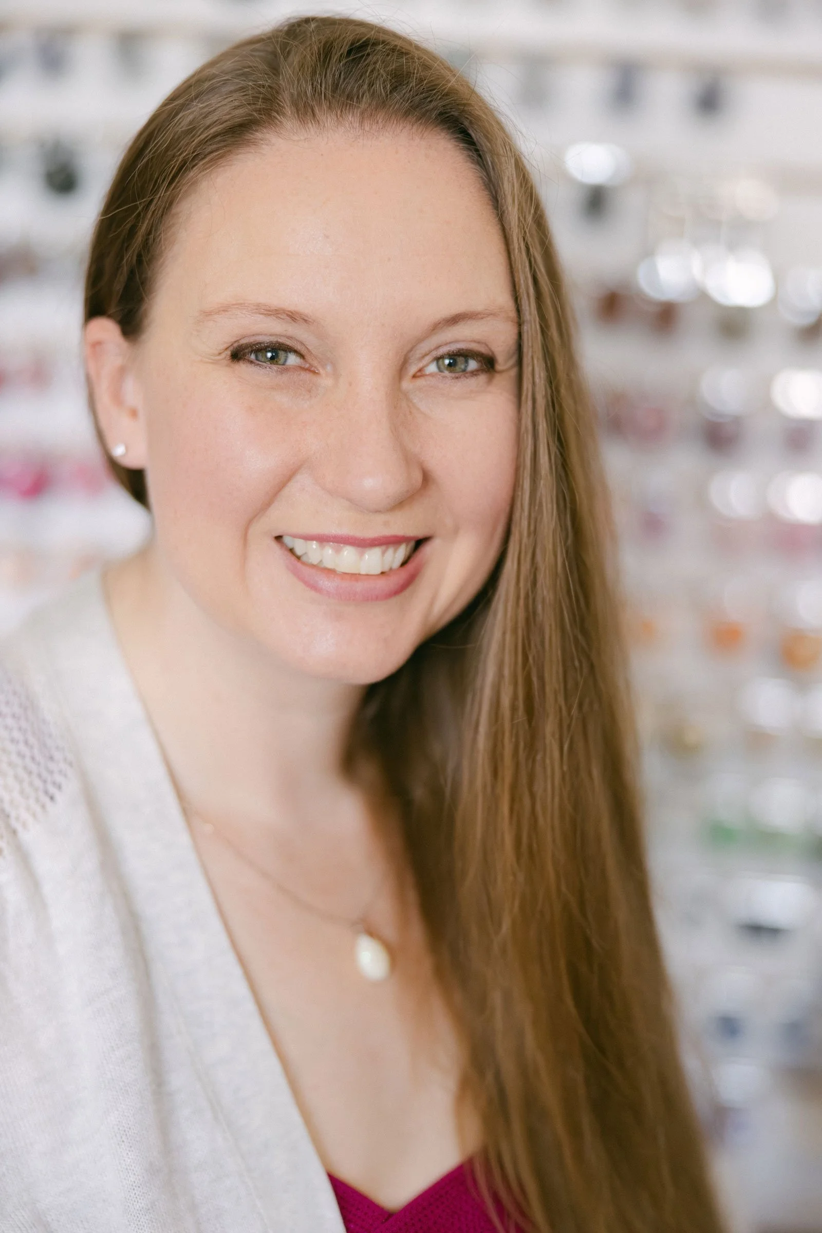 Close-up of a woman with long red hair, light eyes, smiling, wearing earrings and a white top, background with blurry lights.