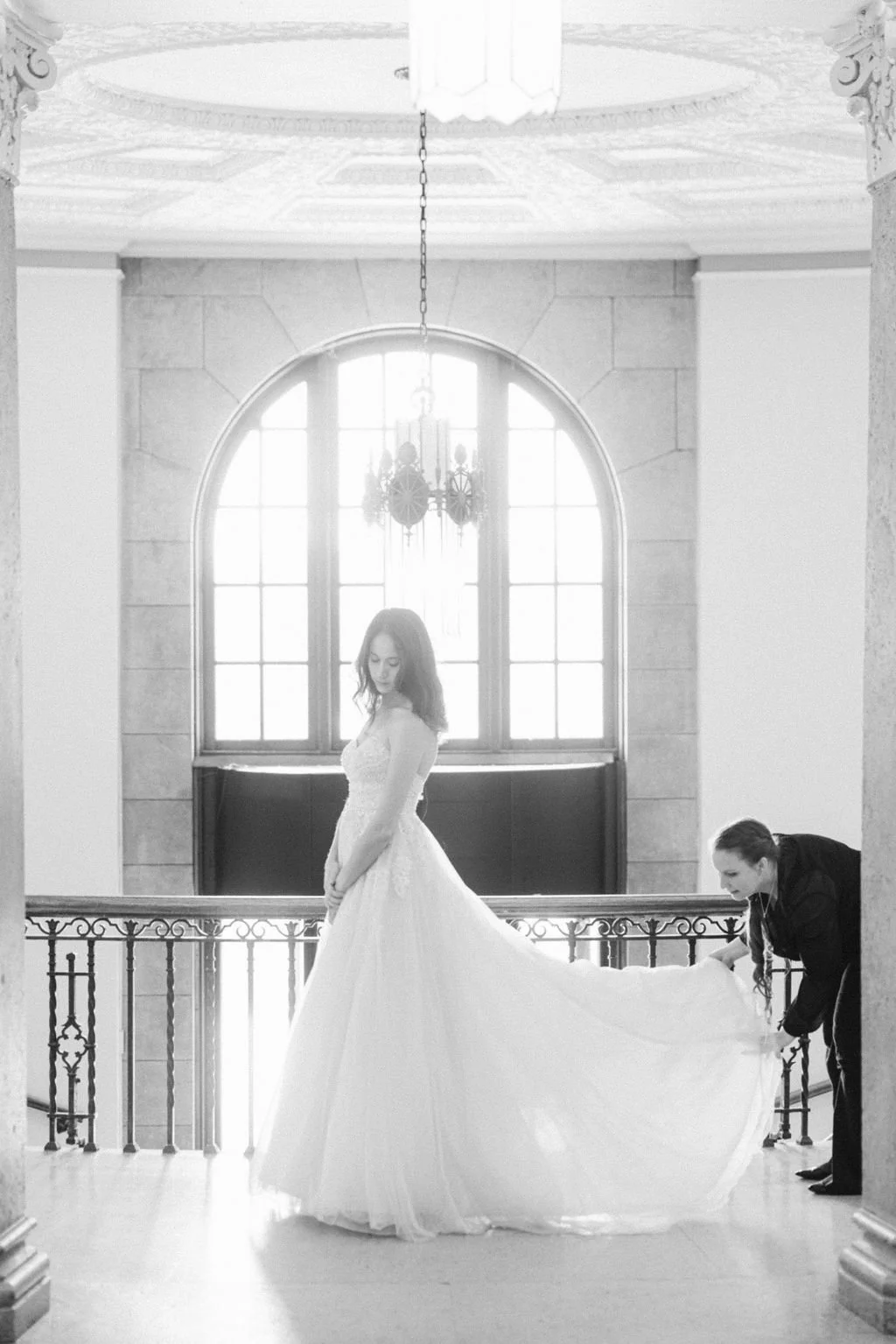 A woman in a wedding dress standing on a balcony with a wedding gown train being adjusted by another woman inside a grand hall with large arched windows and ornate decor.
