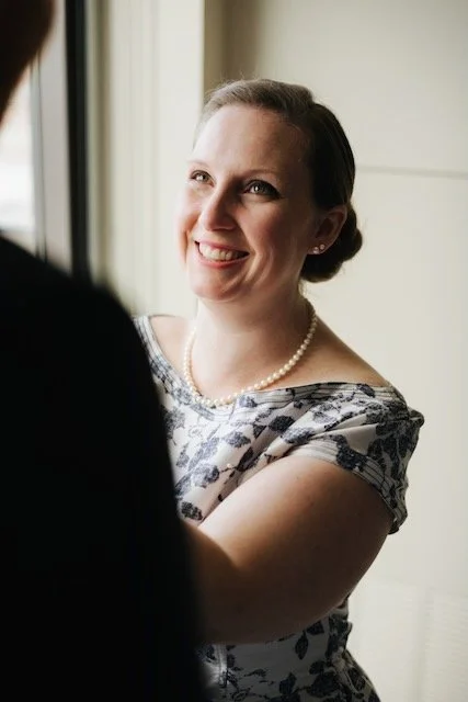 A smiling caucasian woman, Emily, pinning a boutonniere onto a groom