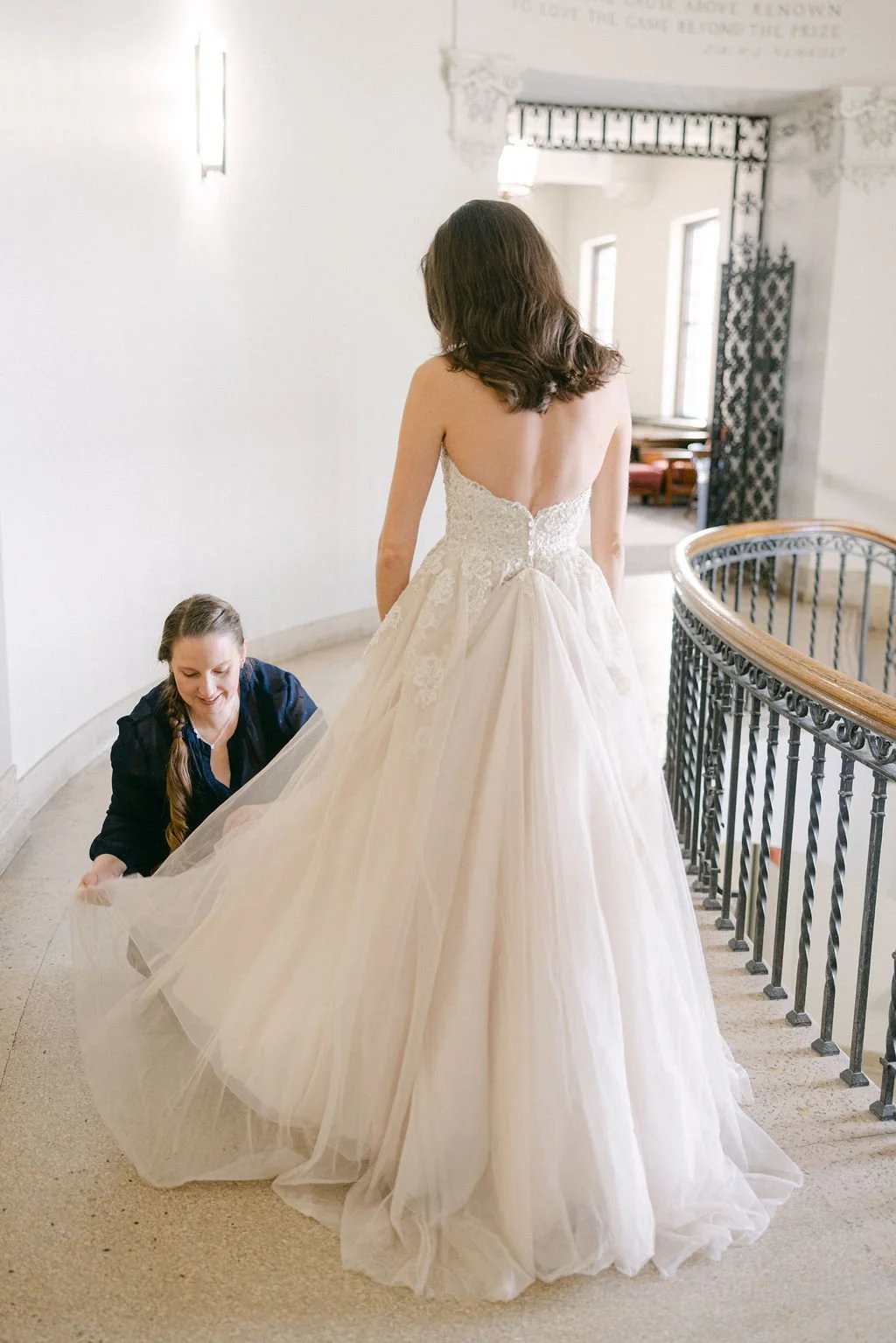 A woman in a wedding dress standing on a staircase while another woman adjusts the train of the dress.