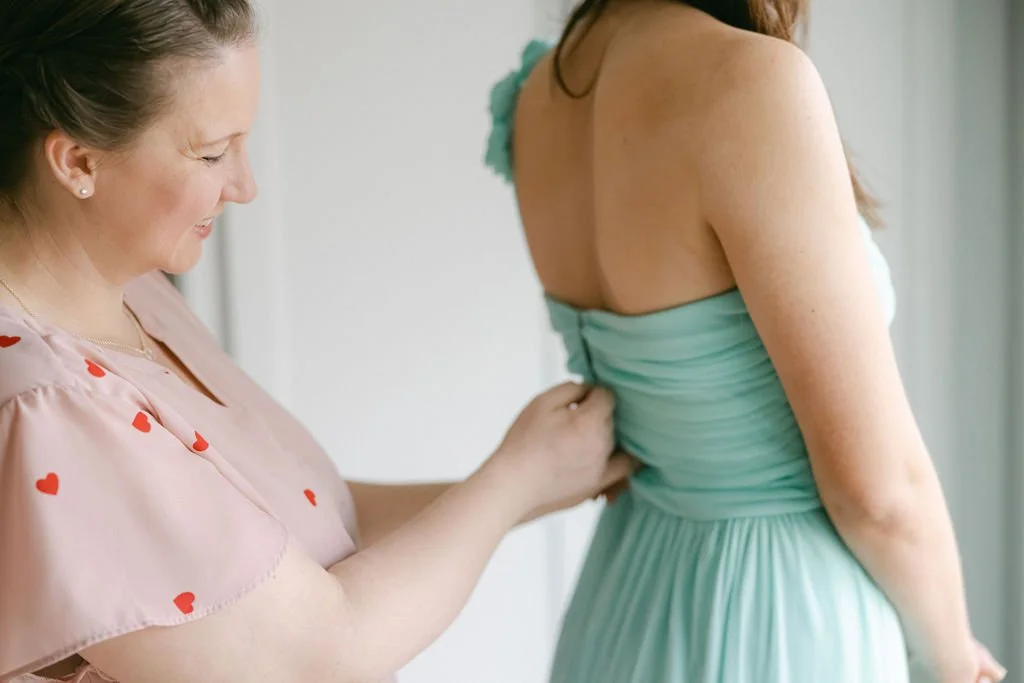 A woman in a pink top with red heart patterns adjusts her dress as she helps another woman in a light green dress.