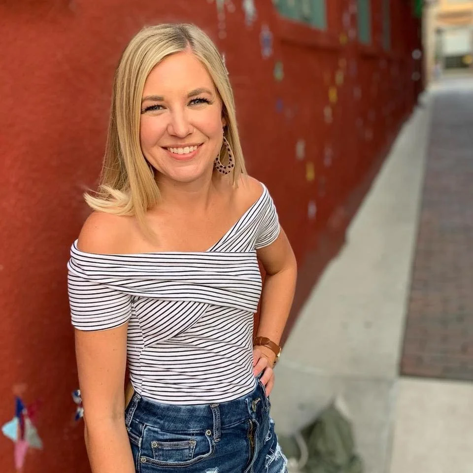 A woman with shoulder-length blonde hair, smiling, wearing a striped off-the-shoulder top and denim shorts, standing against a red brick wall.