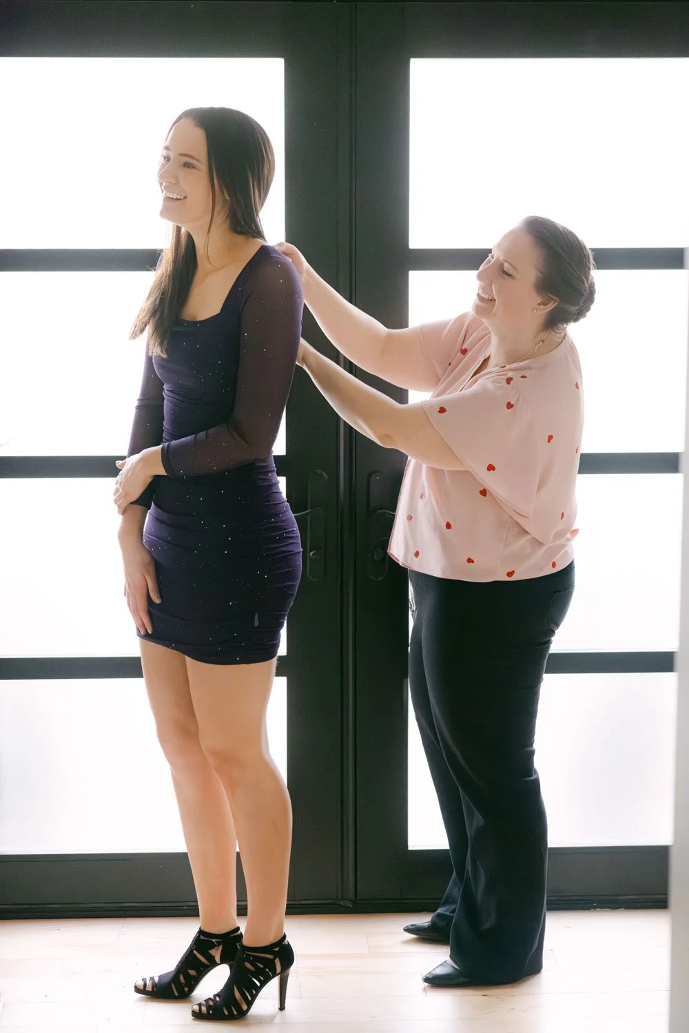 A woman in a dark blue, fitted dress stands in front of a large window as another woman adjusts her dress. Both are smiling and laughing.