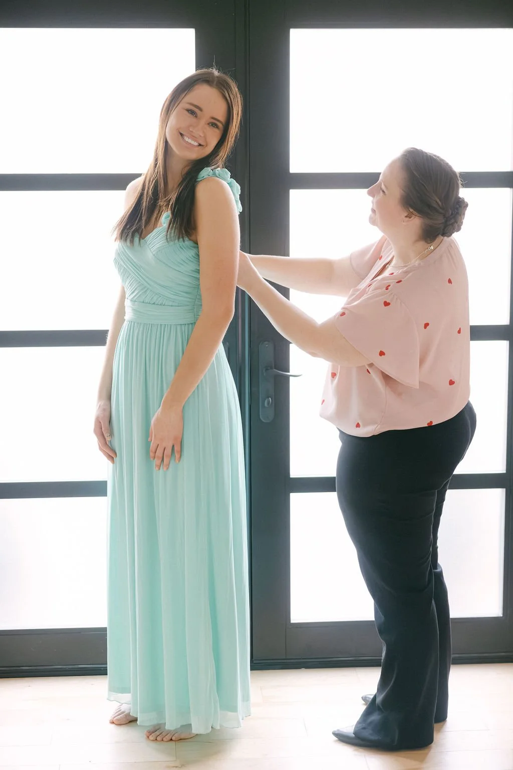 A woman in a pastel blue dress smiling as another woman helps fasten the dress in front of large glass doors.
