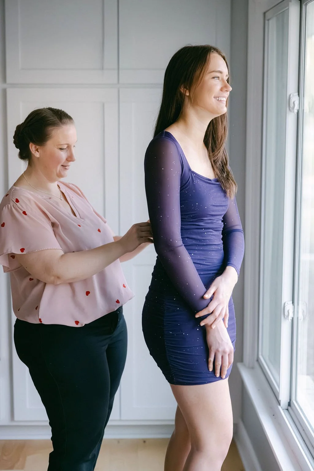 A woman in a blue dress with star-like patterns being helped with her dress by another woman in a pink shirt with small red hearts, near a large window in a bright room.
