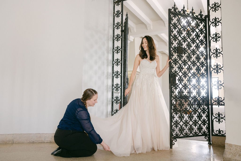 A woman in a wedding dress smiling as another woman adjusts the train of her dress in front of decorative black iron gates.