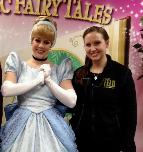 Two women standing together, one dressed as Cinderella in a silver-blue gown with puffy sleeves, gloves, and a headband, and the other in casual black clothing with a badge that says 'FELD', in front of a colorful fairy tale-themed backdrop.