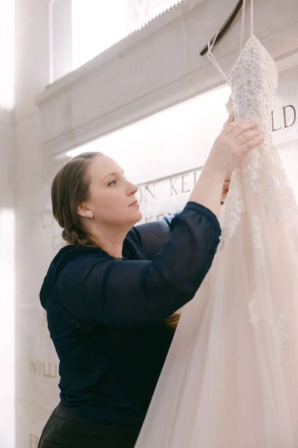 A woman with brown hair pulled back, wearing a dark blouse, is examining a wedding dress on a display. The dress is white with lace and bead details, hanging on a wall in a bridal store.