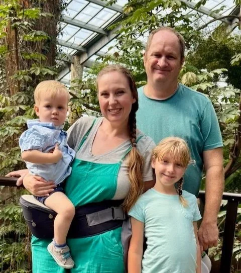 A family of four standing inside a greenhouse surrounded by green plants. The mother is holding a young boy, and a young girl stands beside her. The father stands behind them, smiling.