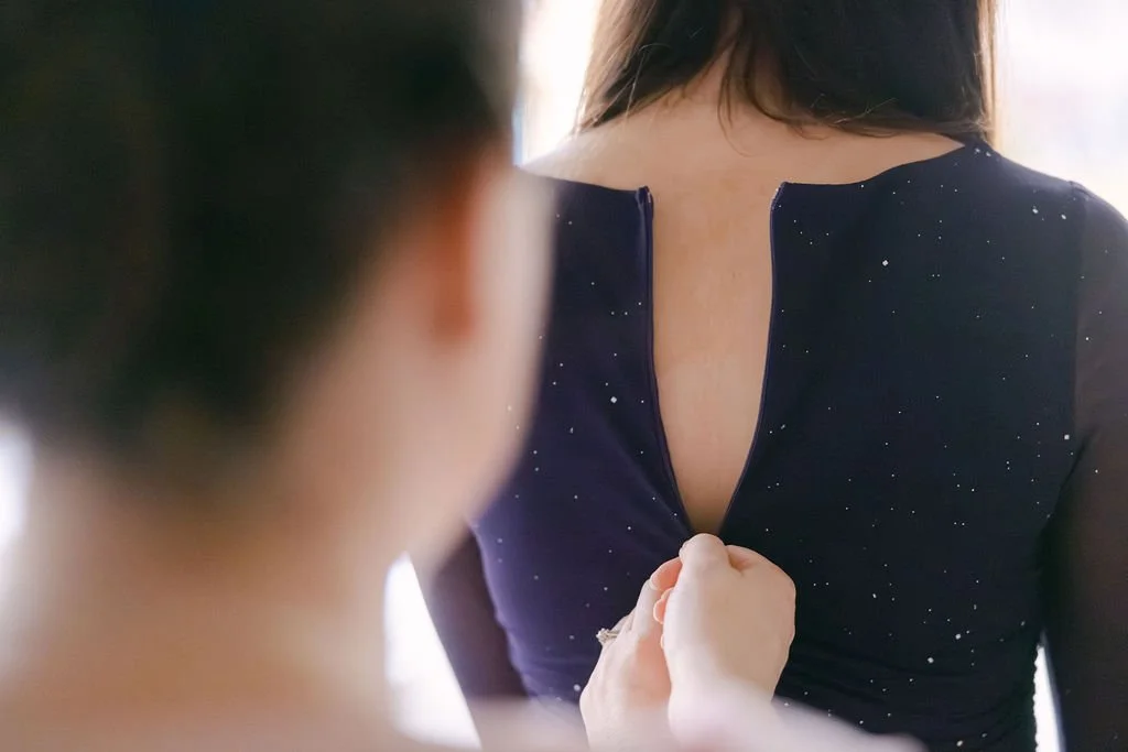 A woman is being helped to zip up the back of her dark blue dress with small white specks.