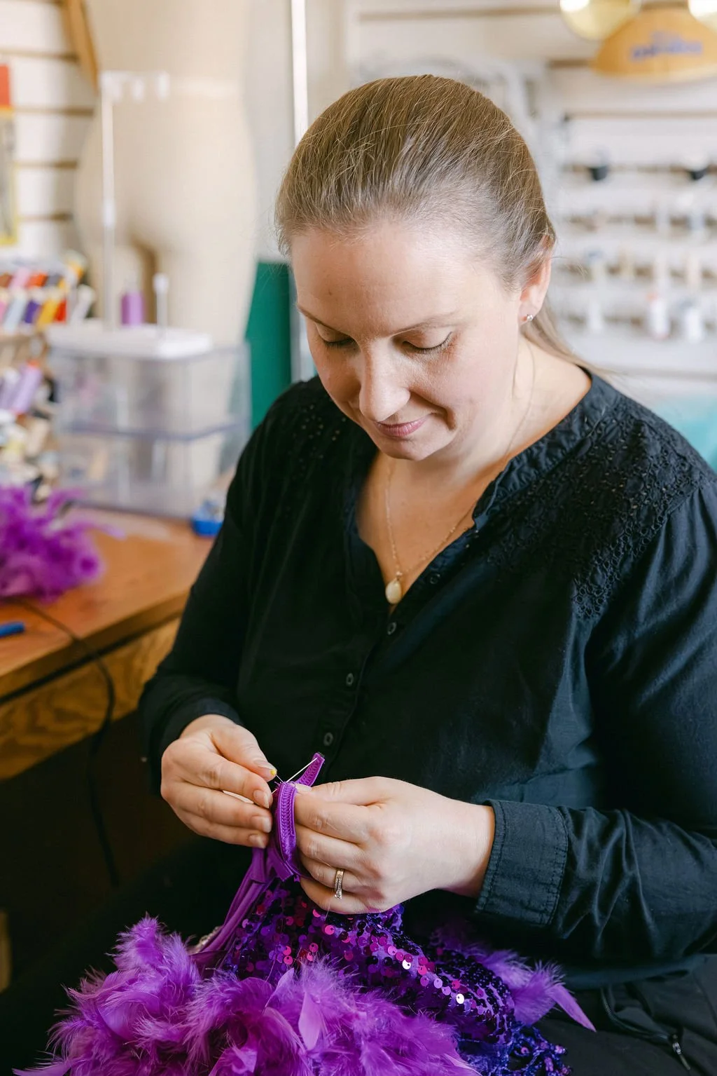 A woman sewing purple feathers onto a sequined fabric in a craft room.