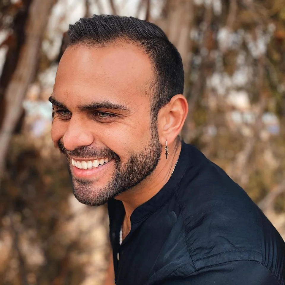 A smiling man with a beard, wearing a black shirt and an earring with a cross, outdoors with blurred trees in the background.