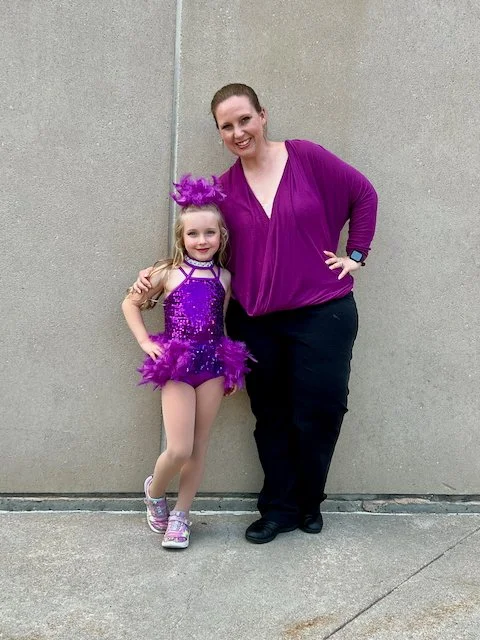A young girl in a purple dance costume with a tutu and glitter, standing next to an adult woman in a purple top and black pants, both smiling against a plain wall.