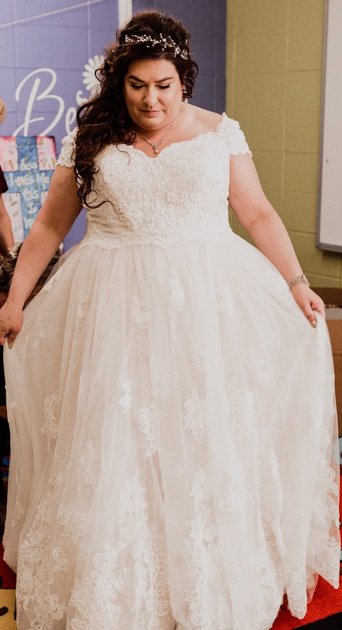 A woman in a white wedding gown with lace details, holding the skirt out slightly, standing in a room with a purple wall and a decorated background.